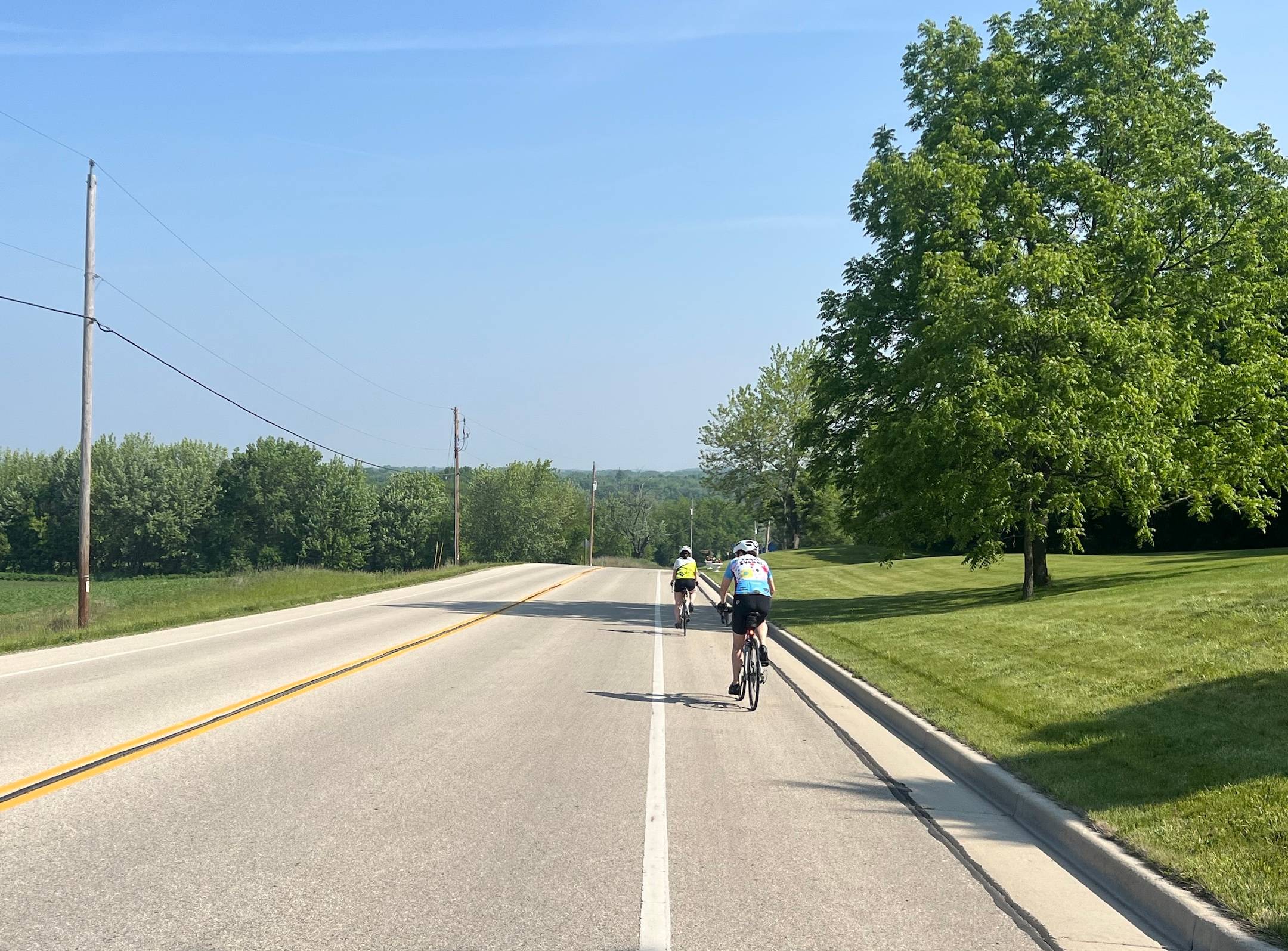 Two cyclists ride the shoulder of a two-lane road through a hilly scene of lawns and trees.
