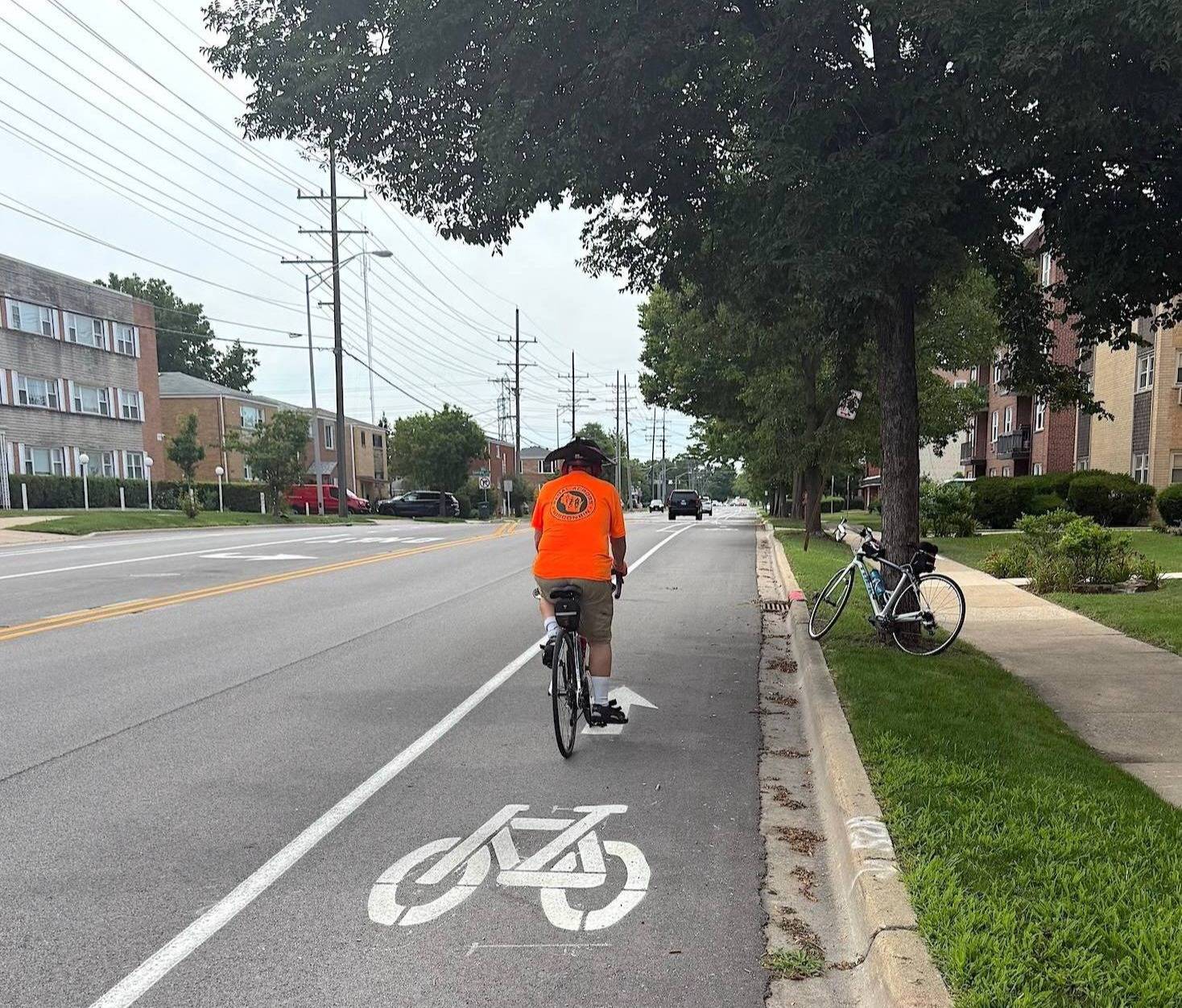 A casual bicyclist in a bright orange T-shirt and sun-protecting hat rides in a bike lane on a newly paved suburban road under short trees.