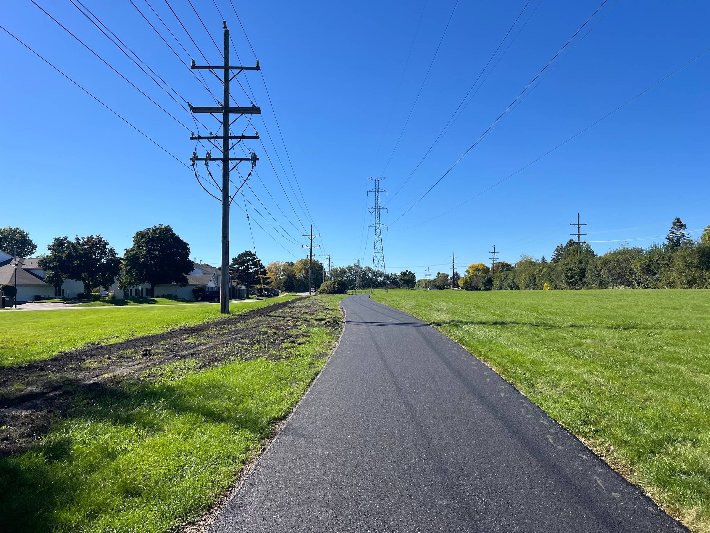 A very newly asphalt-paved trail extends ahead in an open field with power lines overhead, with suburban homes in the background on the left