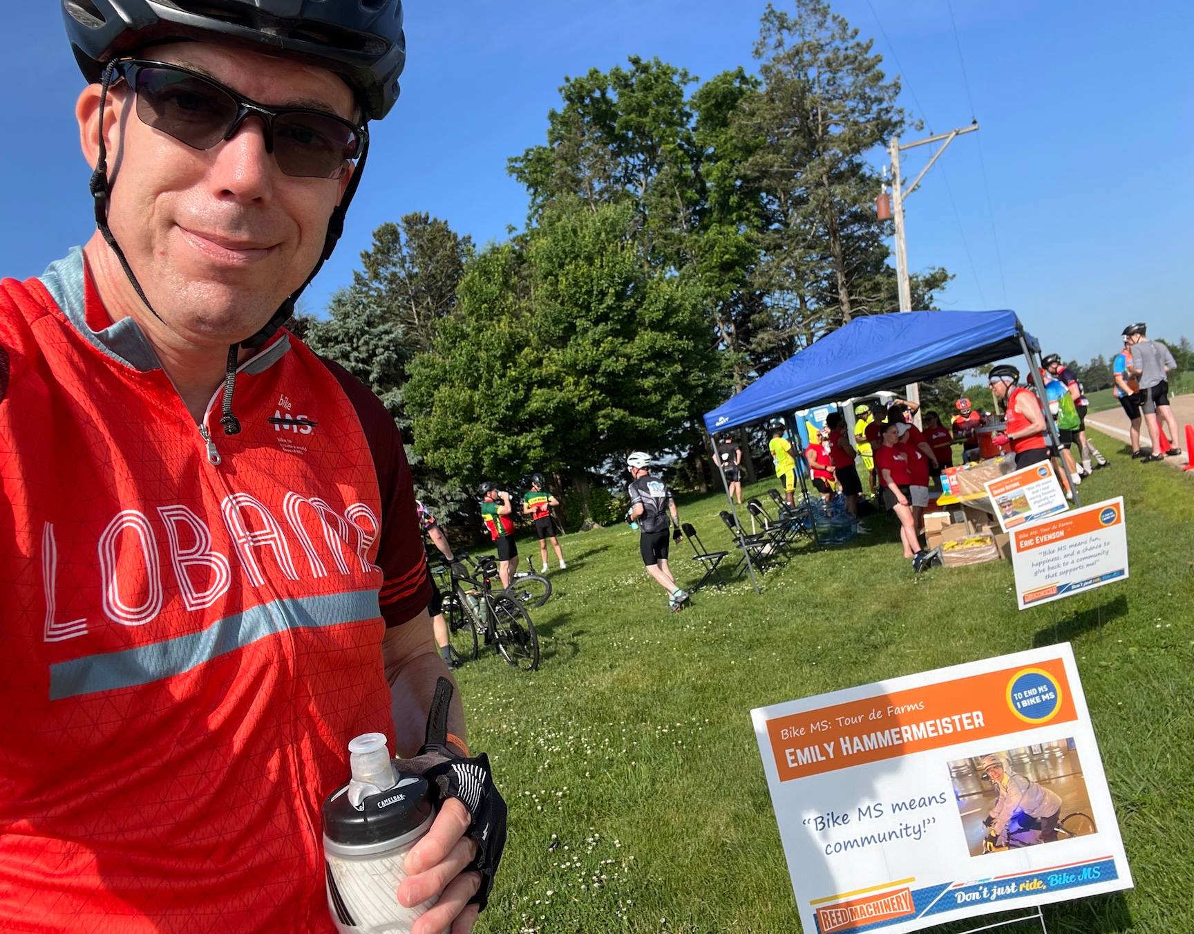 Neil, in a helmet, sunglasses and an orange bike jersey, and holding a water bottle, takes a selfie in front of a picnic area of a group ride.