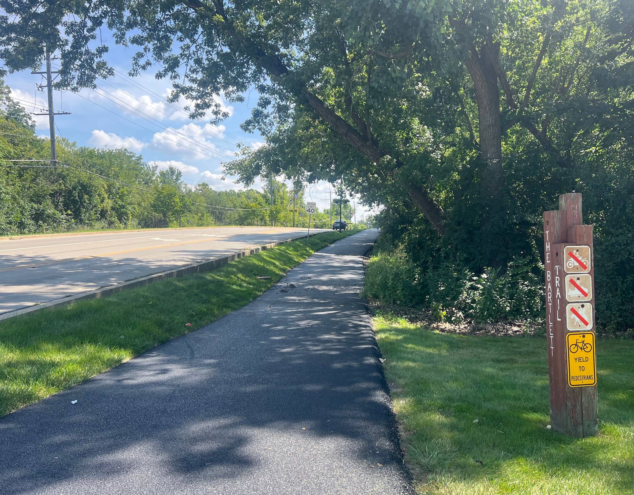 A new black asphalt path runs by "The Bartlett Trail" signpost with other bike signs on the right, under trees' shade and along a tree-lined street.