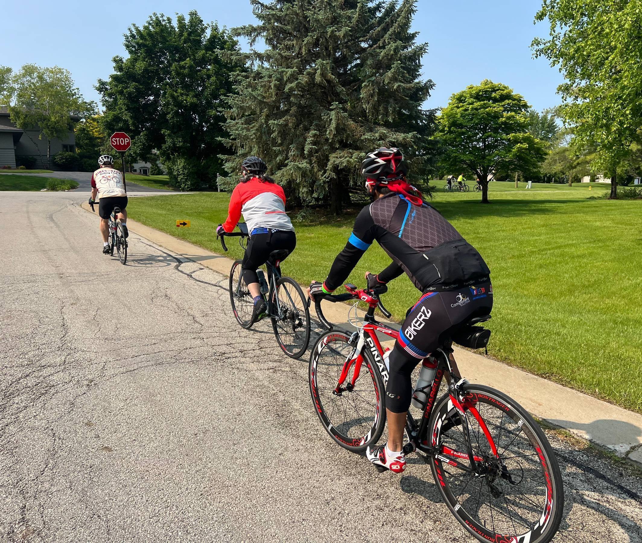 A few bicyclists ride on a suburban residential street with lawn and trees in the background.