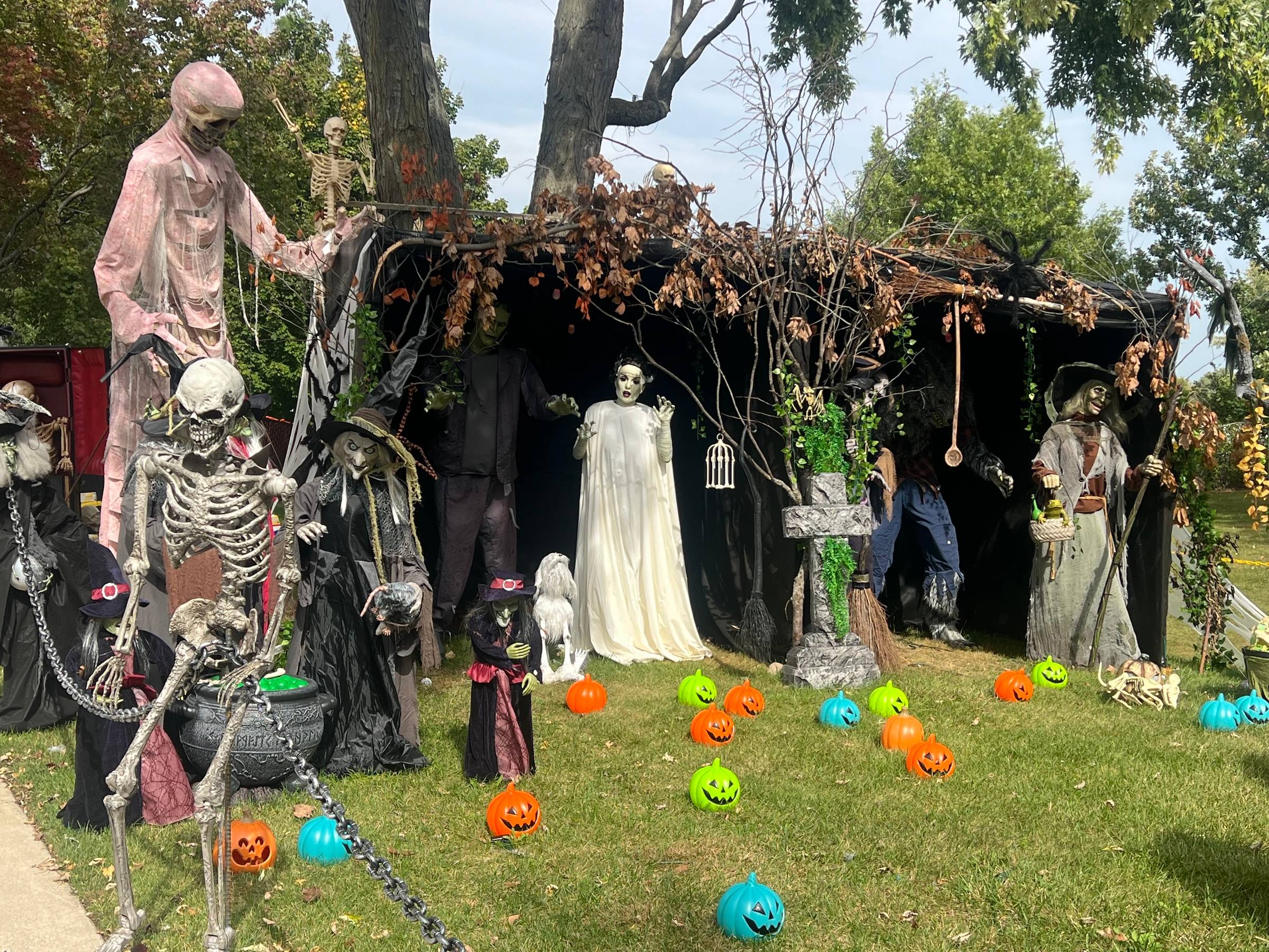A scene full of Halloween creatures tall and short, with the bride of Frankenstein in the center, and colored fake pumpkins decorate a lawn under trees.