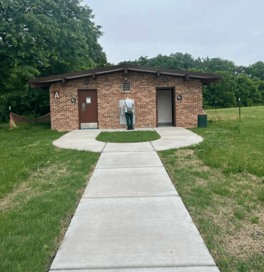 A newly paved sidewalk leads to a forest preserve restroom building with two doors and a drinking fountain in the middle.