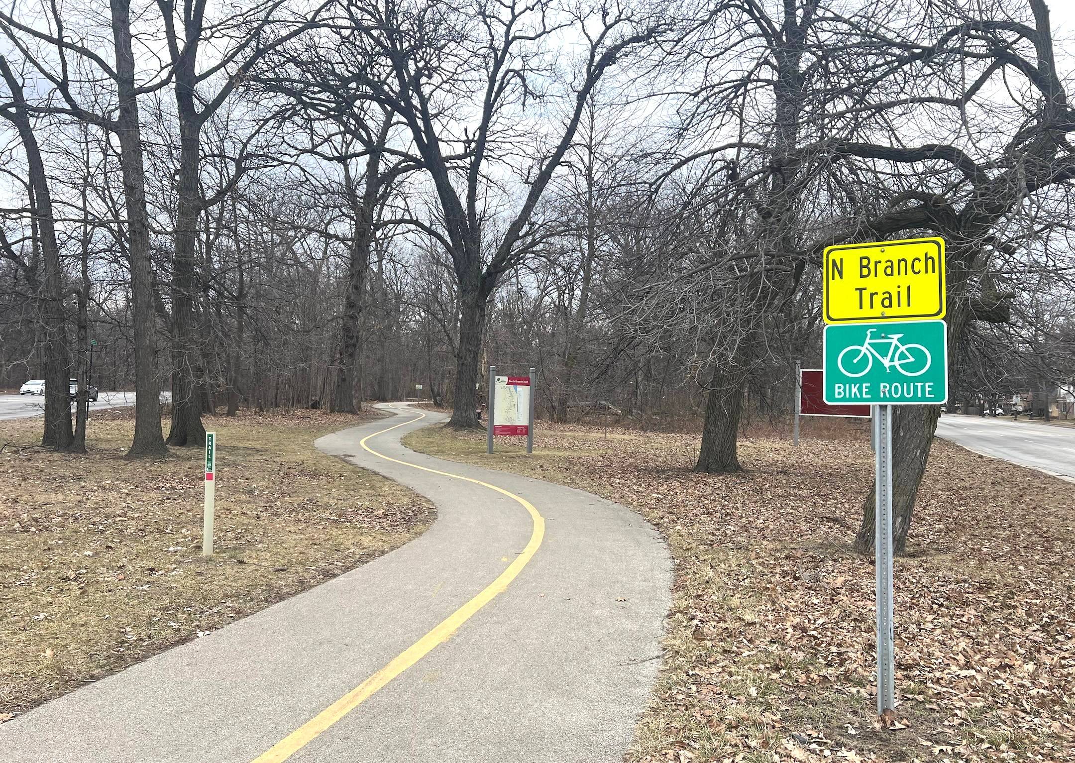 Looking down a curved, striped forest preserve bike trail with bike route signage and bare trees in winter