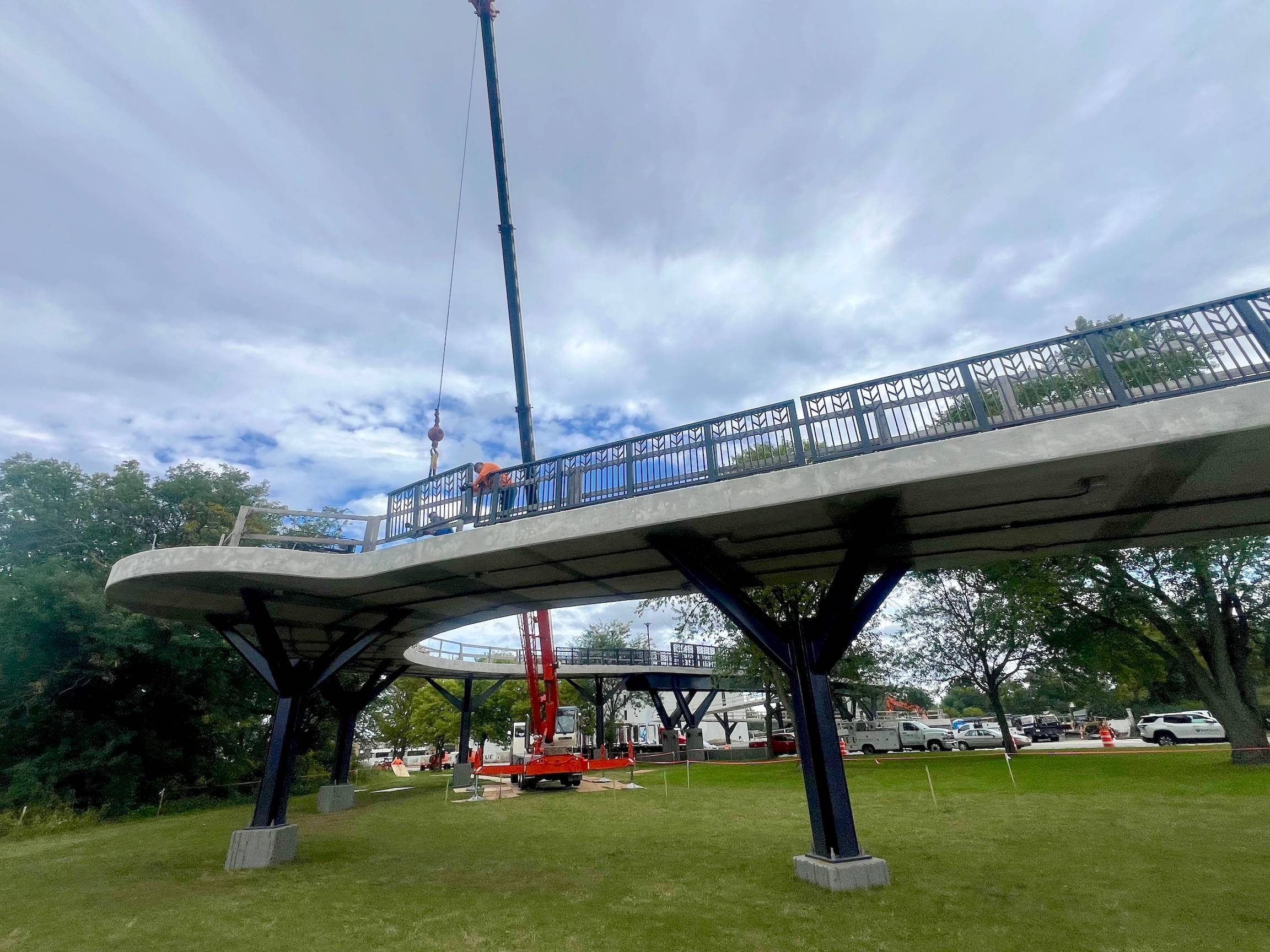 Workers place a railing hung off a crane into place on a new cement bridge in a forest preserve by a suburban street.