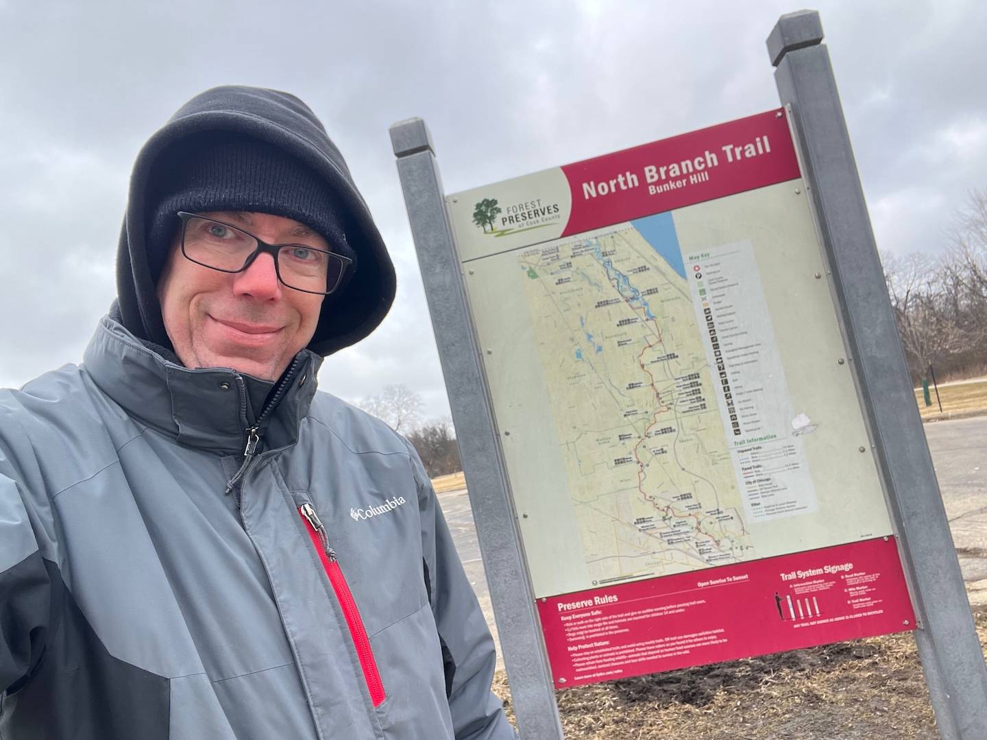 A man in a winter coat, ski cap, hood and glasses stands in front of a forest preserve trail map sign.