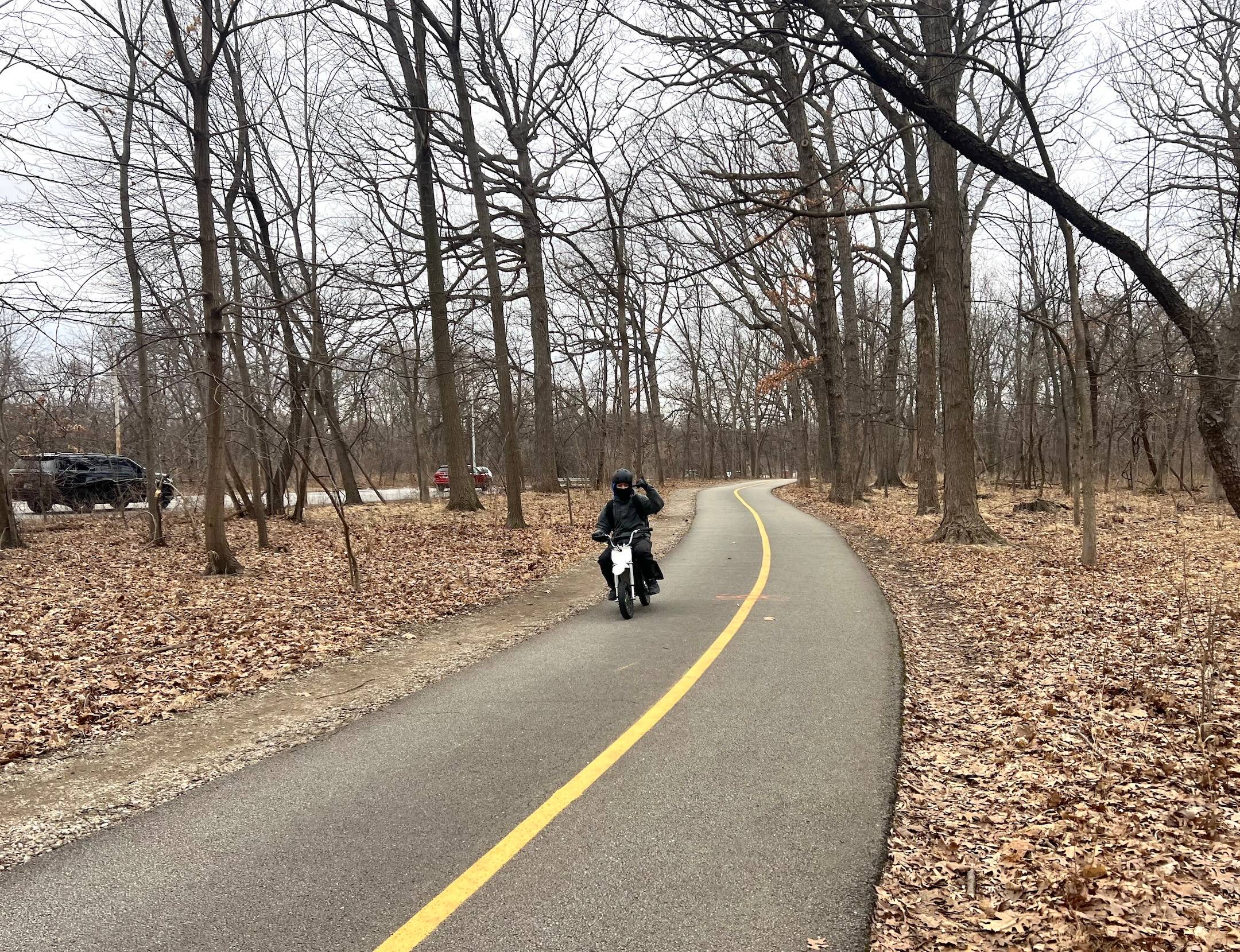 A bundled-up rider of a small motorbike makes a devil's-horn finger gesture as he zips down a striped bike trail through a forest preserve.