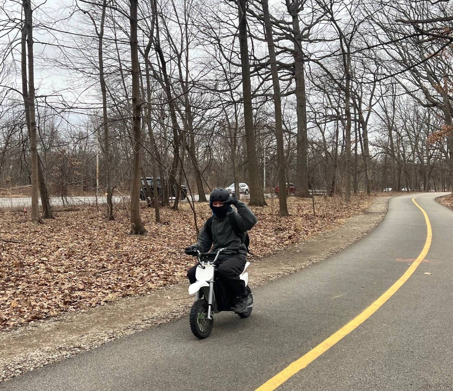 An e-moto rider bundled up in black flashes fingers as he comes down a striped bike trail in a forest.