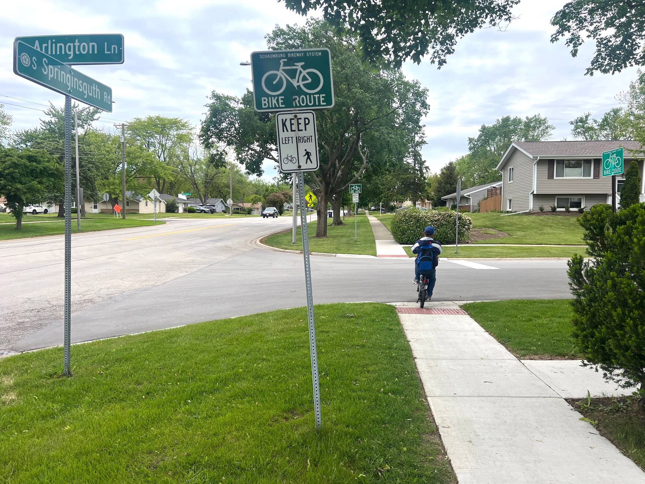 A bicyclist on a short e-bike rides on a neighborhood sidewalk and crosses a street, with Bike Route and other signs up front.