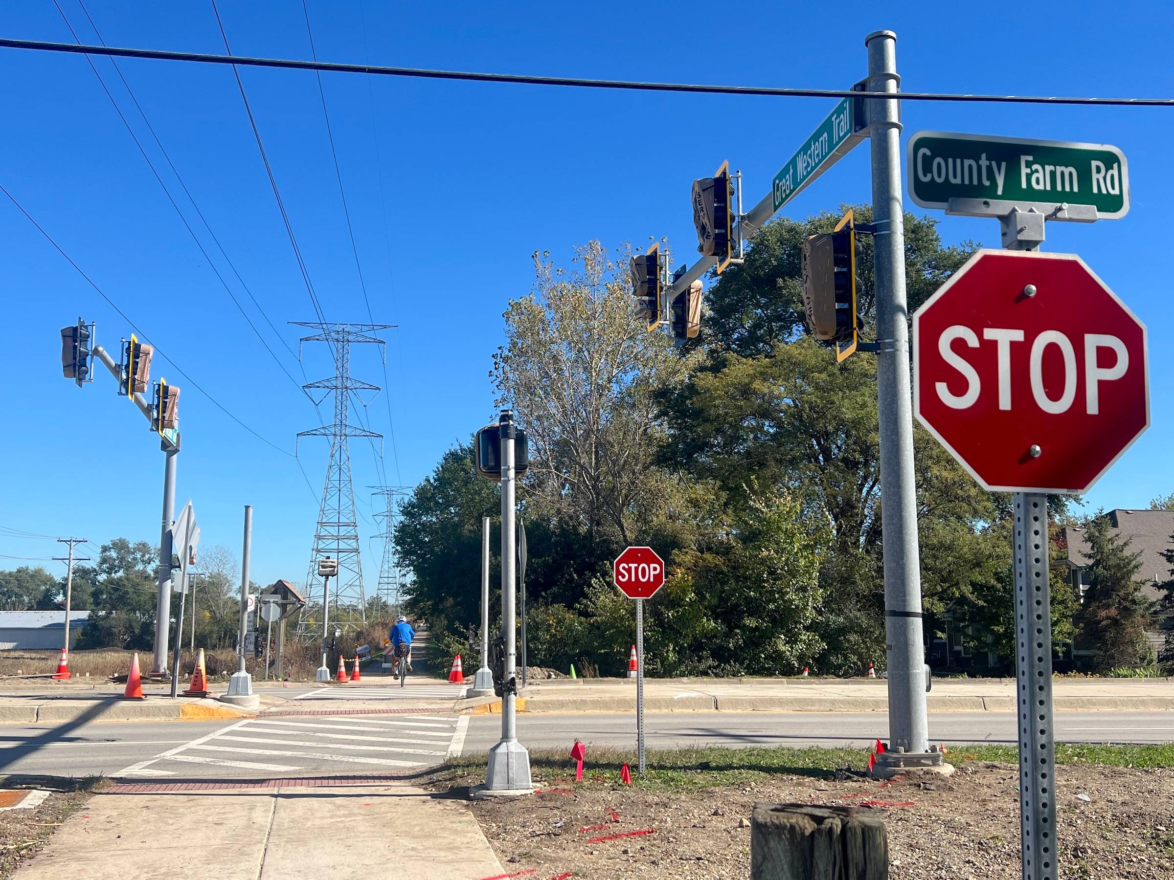 Two Stop signs appear at a bike trail crossing with a busy suburban road that has a median and new, still-covered traffic signals, trees and power lines in the background