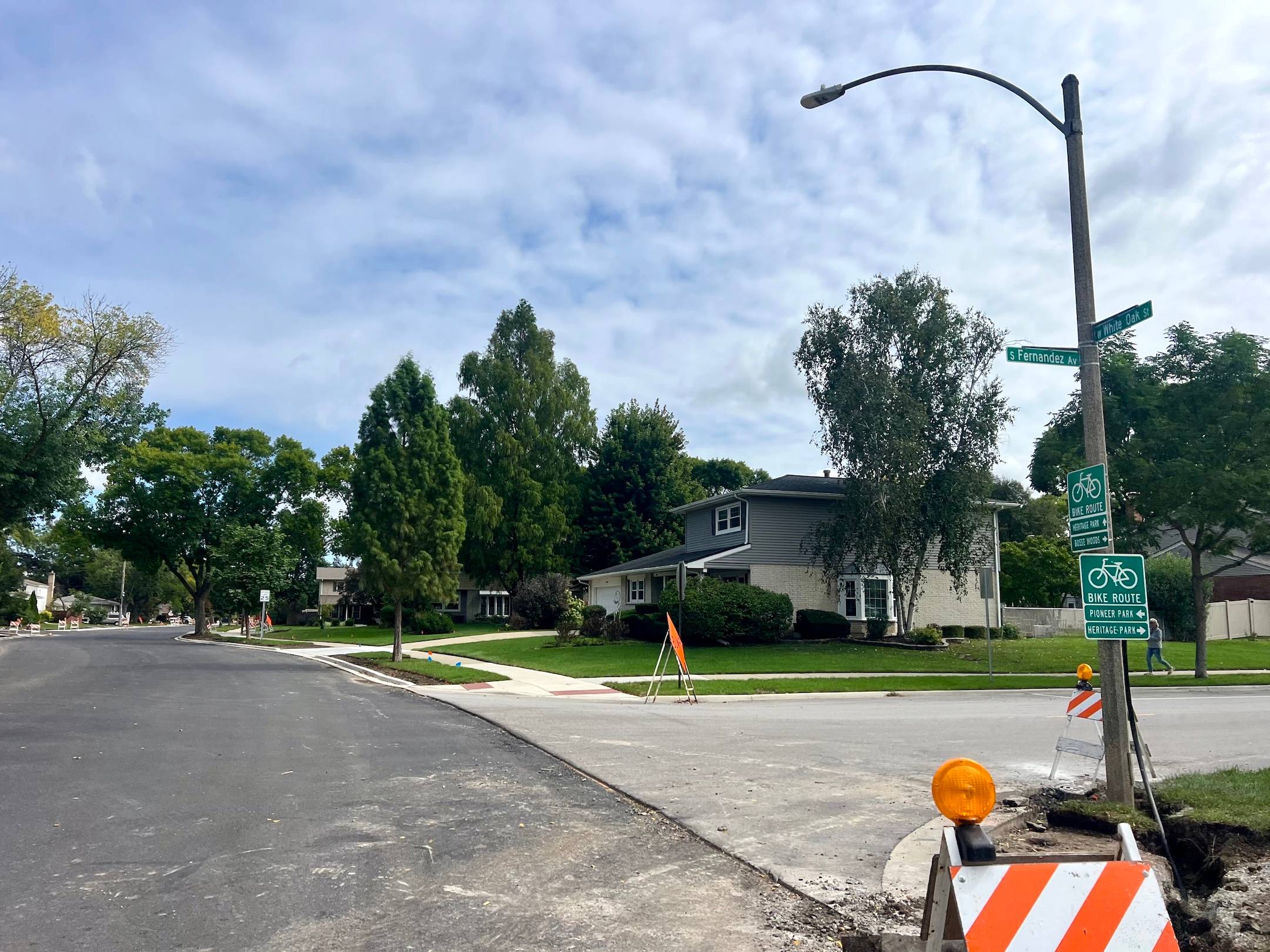 A first layer of asphalt appears on a street in a suburban neighborhood at an intersection with a light pole and bike route signs, with a construction horse up front.