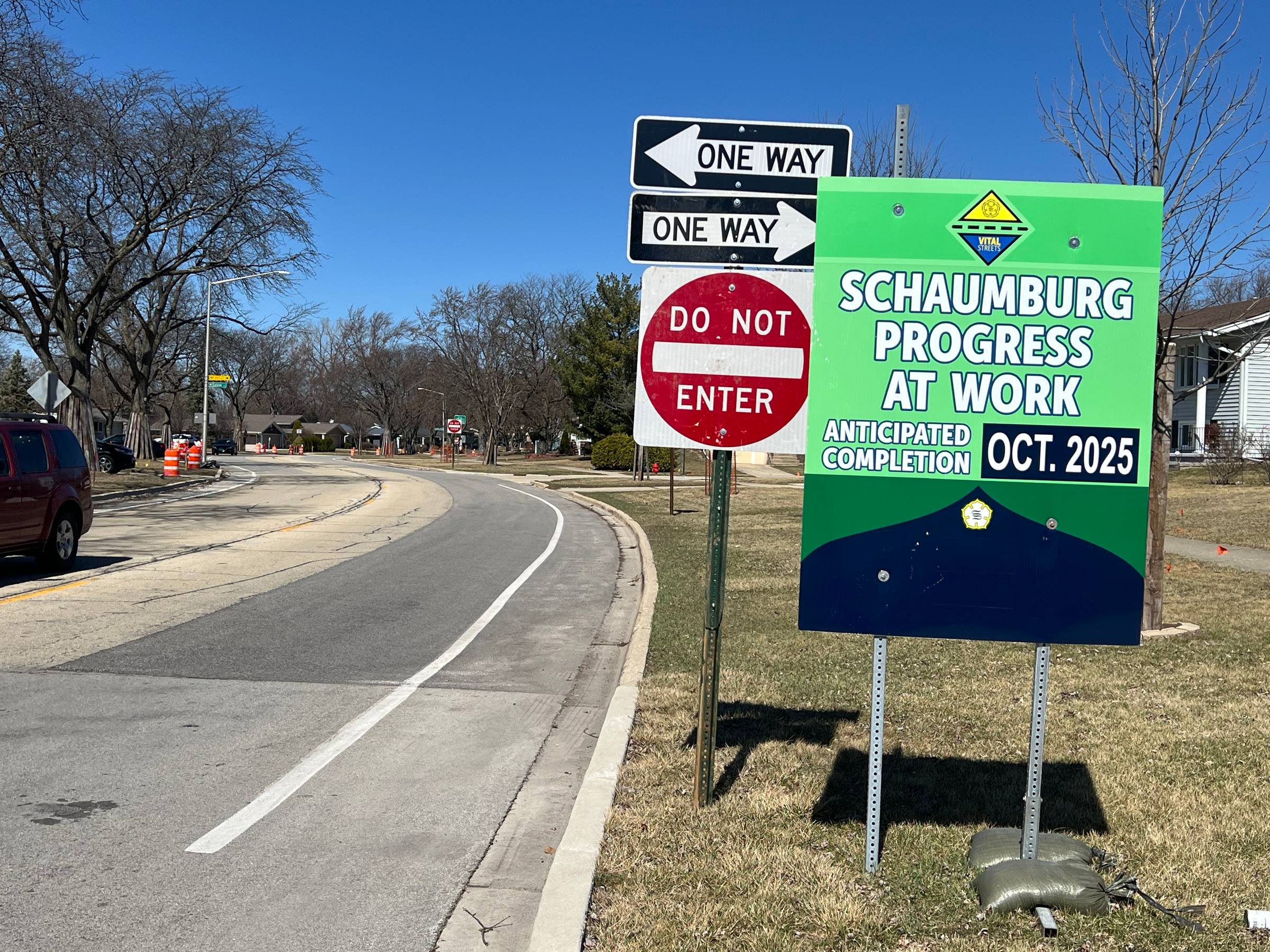 "Schaumburg Progress at Work," two One Way signs pointing in opposite directions, and a Do Not Enter sign appear alongside a paved suburban neighborhood road with a bike lane.