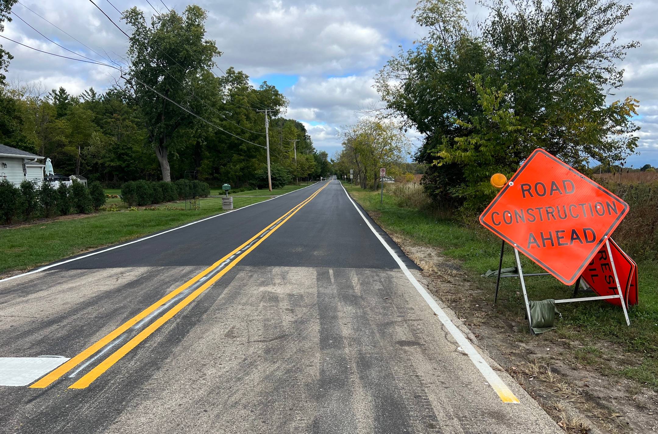 Looking down a newly paved two-lane road with an orange Road Construction sign up front, prairie to the back right, a home behind bushes on the left, and trees lining both sides ahead.