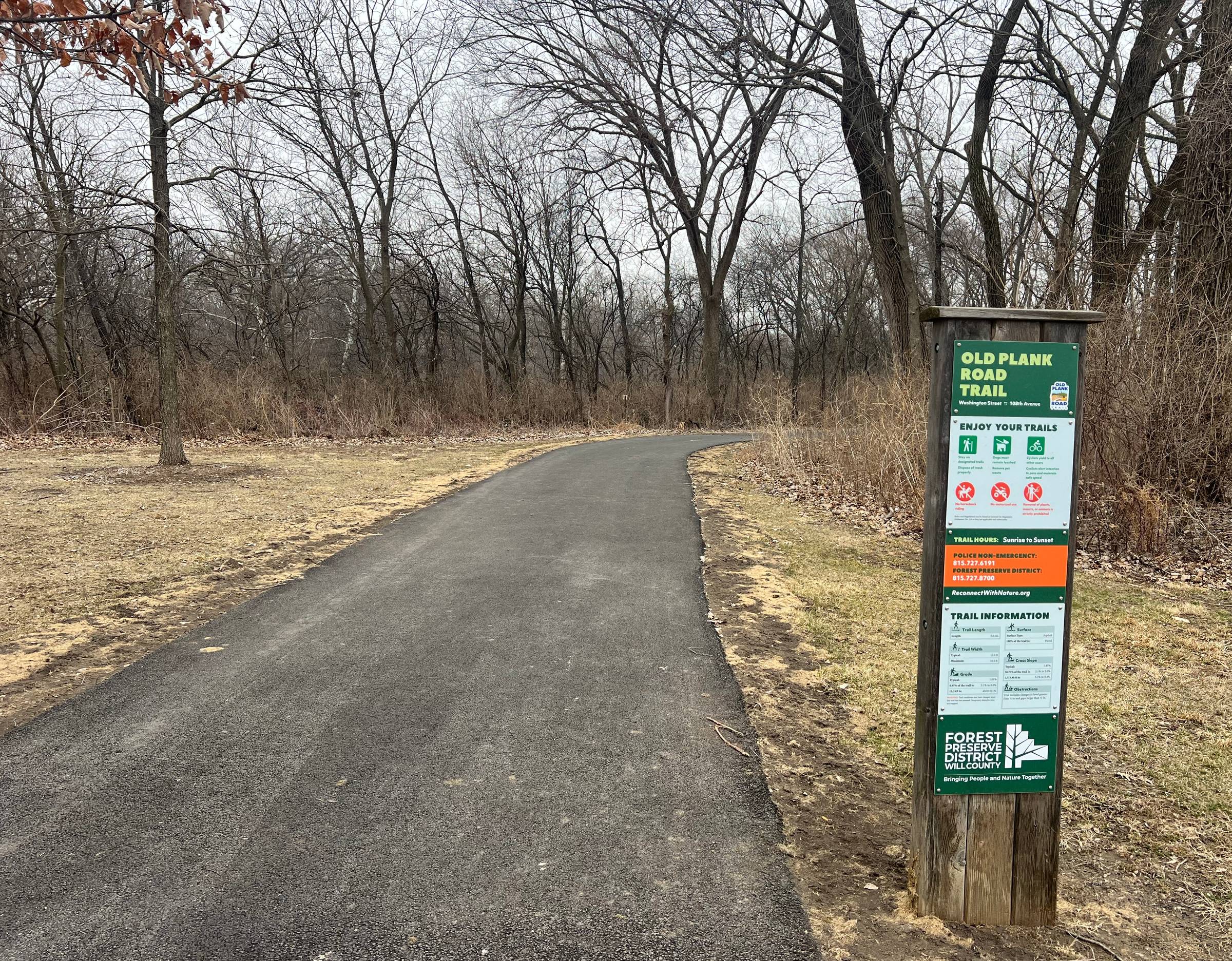 A new black asphalt path proceeds straight and curves right in a forest preserve with signage