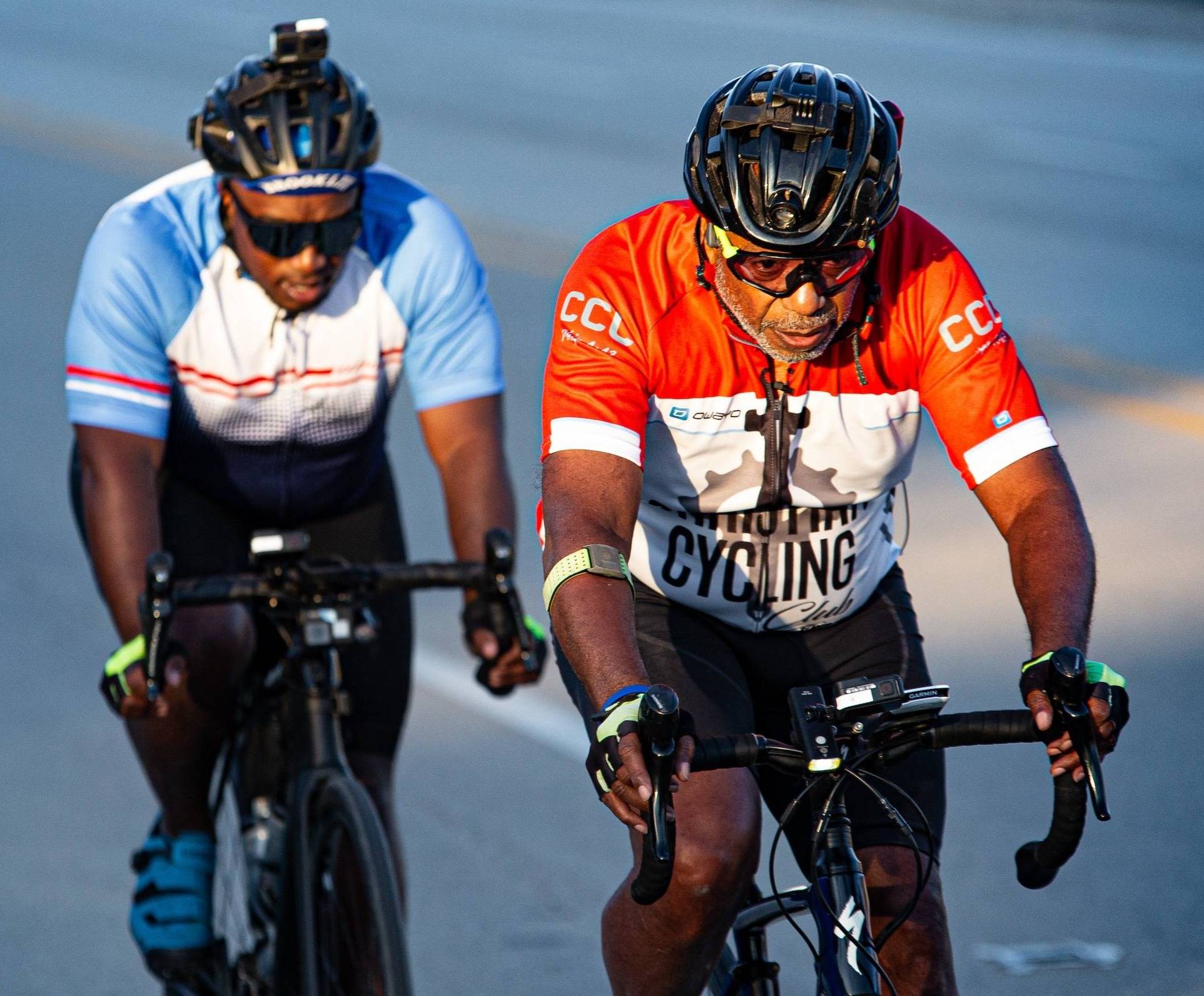 Two cyclists are seen up close, one behind the other, riding road bikes on the shoulder of a road.