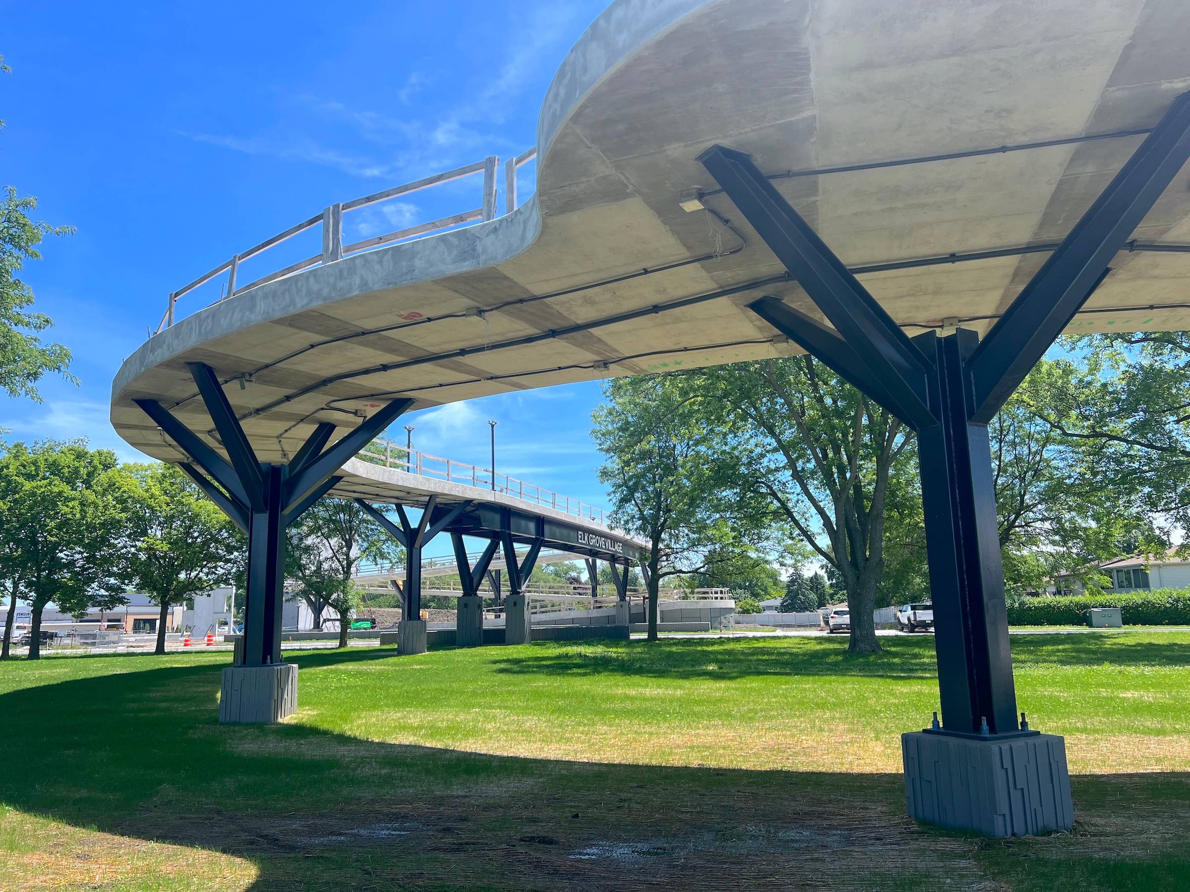 Looking at the underside of a new concrete bike-pedestrian bridge over a lawn with makeshift wooden railings