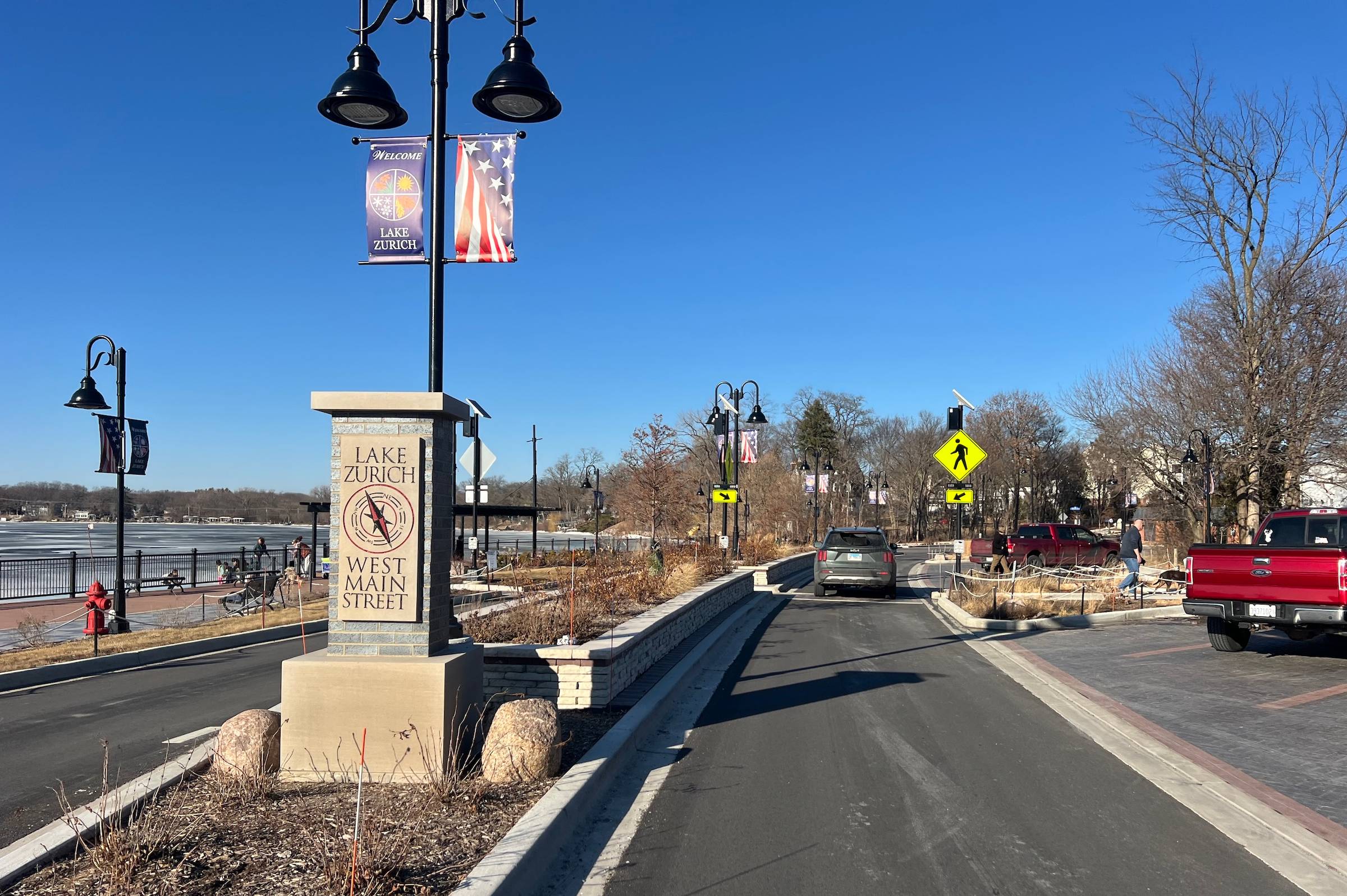 Looking down a newly paved road divided by a big median with a garden, stone signage and streetlamps by an icy lake on the left and colored brick parking spaces on the right