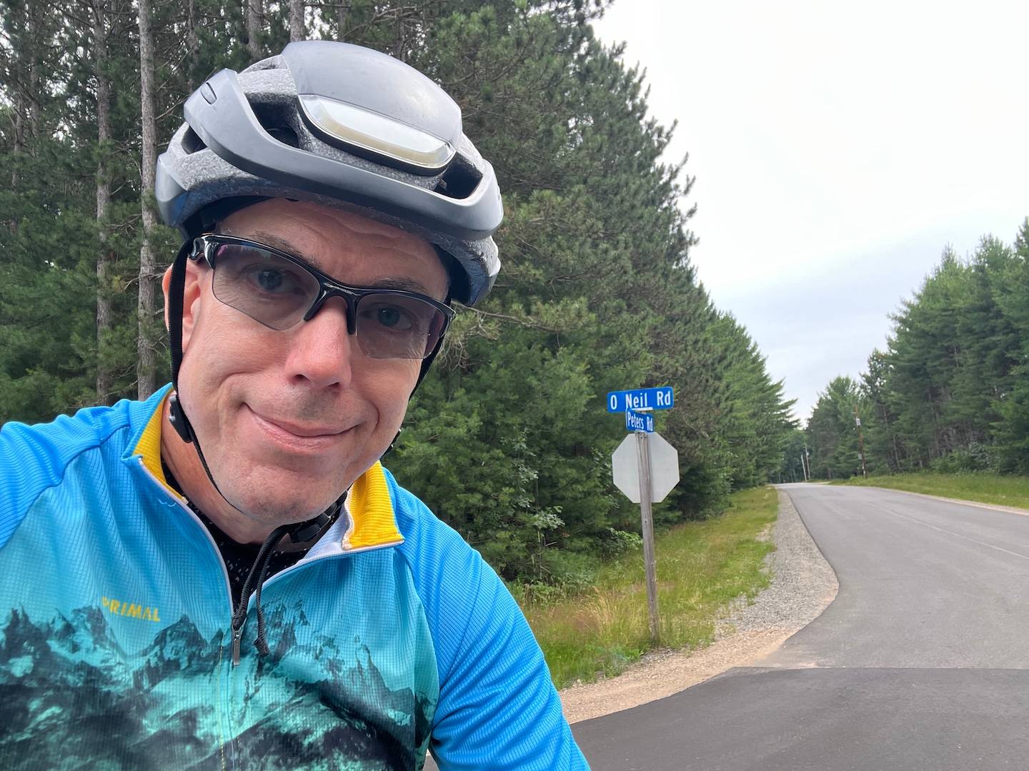 A cyclist in helmet takes a selfie in front of a road lined with tall pine trees under a hazy sky.