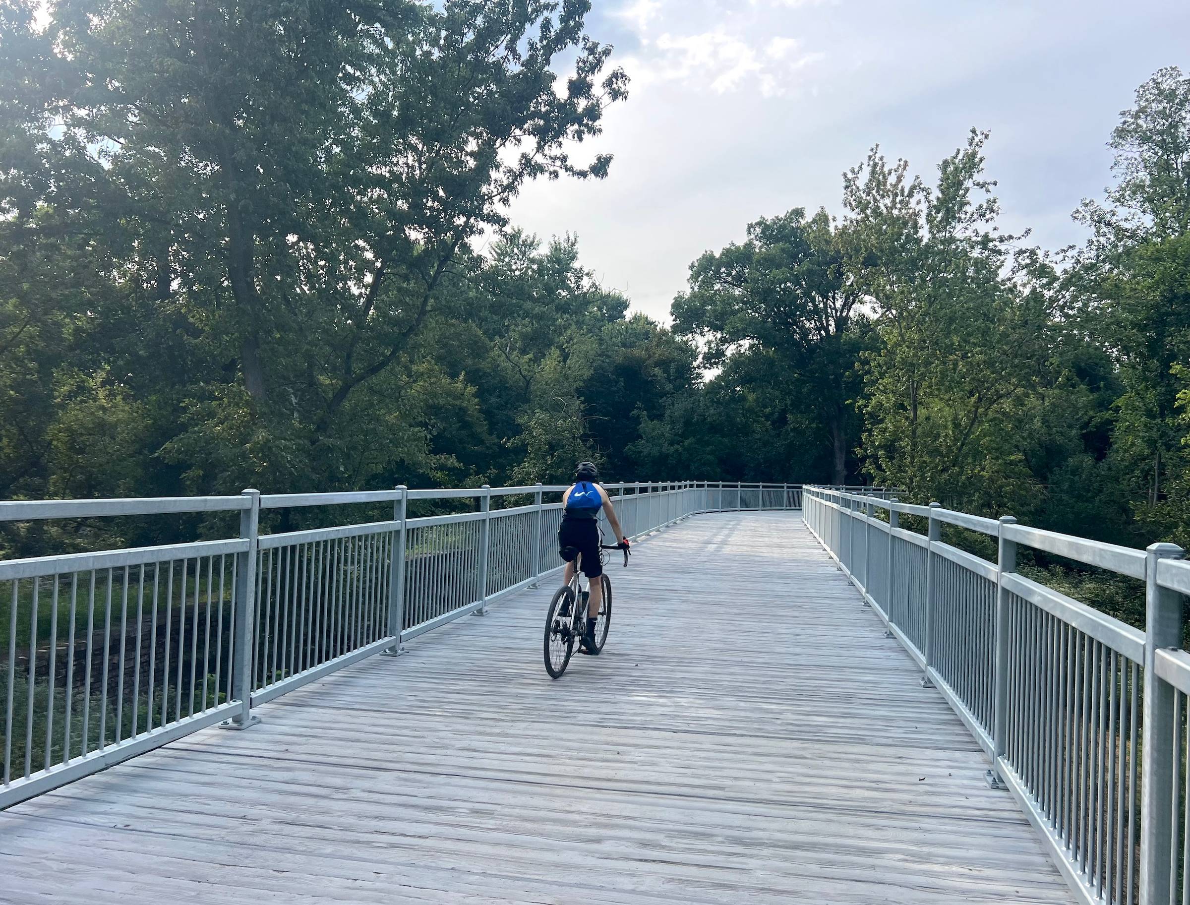 A sleeveless young cyclist with a blue backpack rides on a boardwalk bridge through a forest preserve