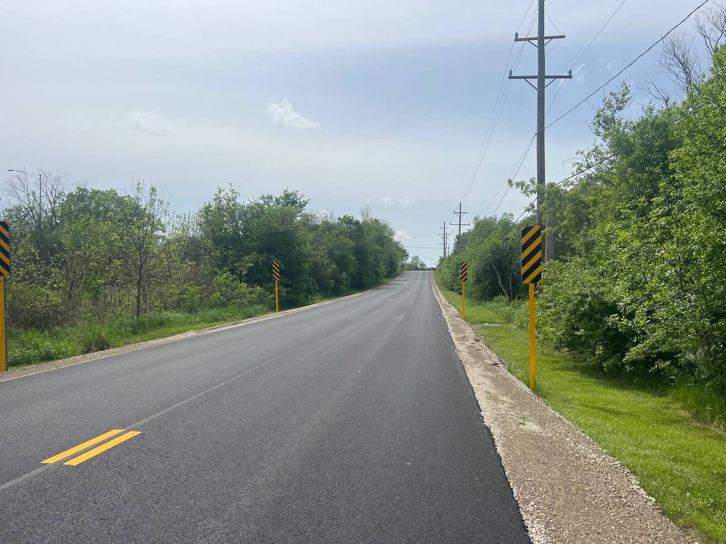 A road through short trees with power lines on the right has new black asphalt and small yellow stripes.