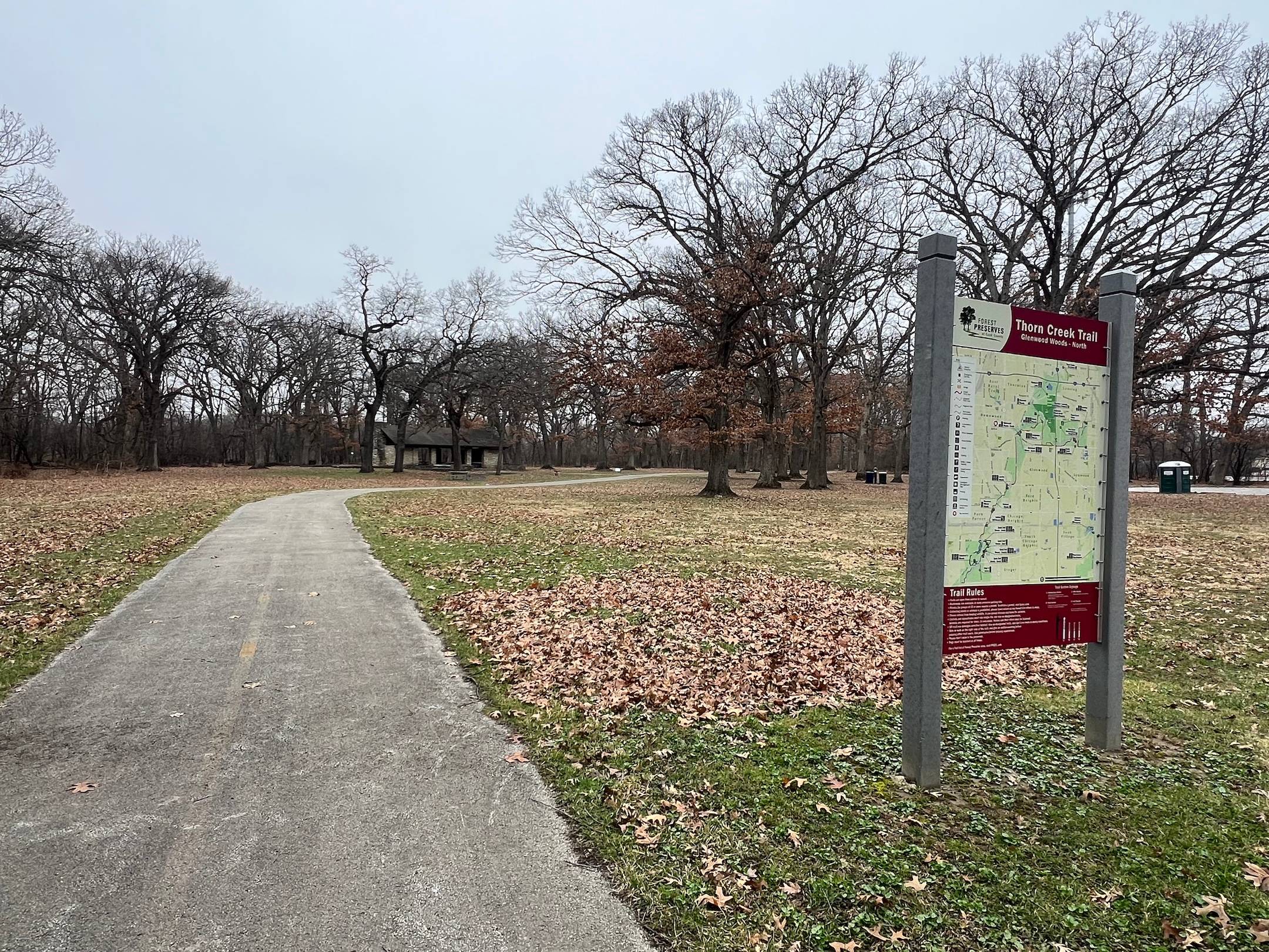 A bike trail with a very faded strip down the middle extends into and curves in a forest preserve with leaves on the ground, not the trees, and a big sign and map on the side.