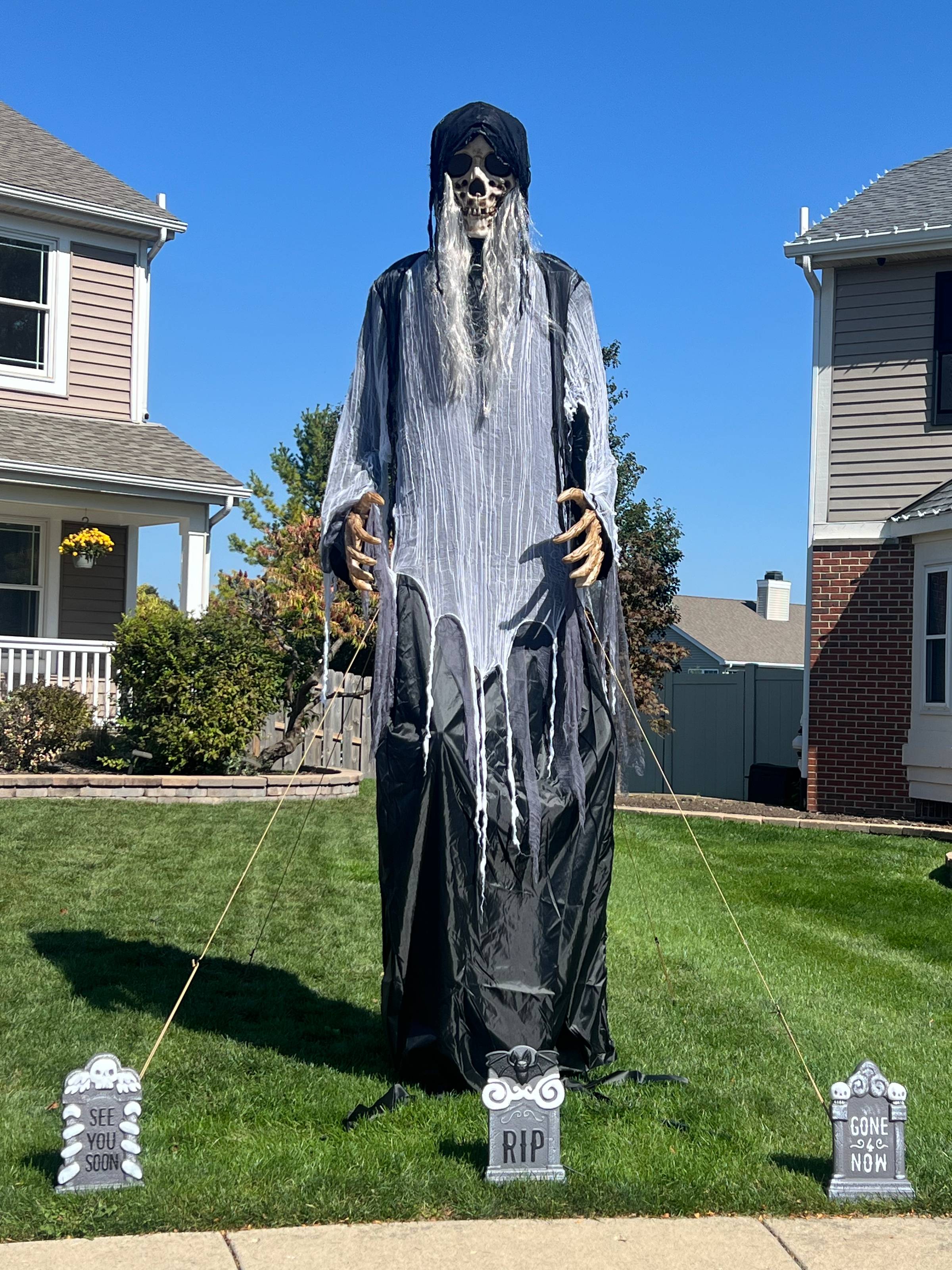 A tall creepy skeleton in a robe with hood and long gray hair stands on a suburban front lawn.