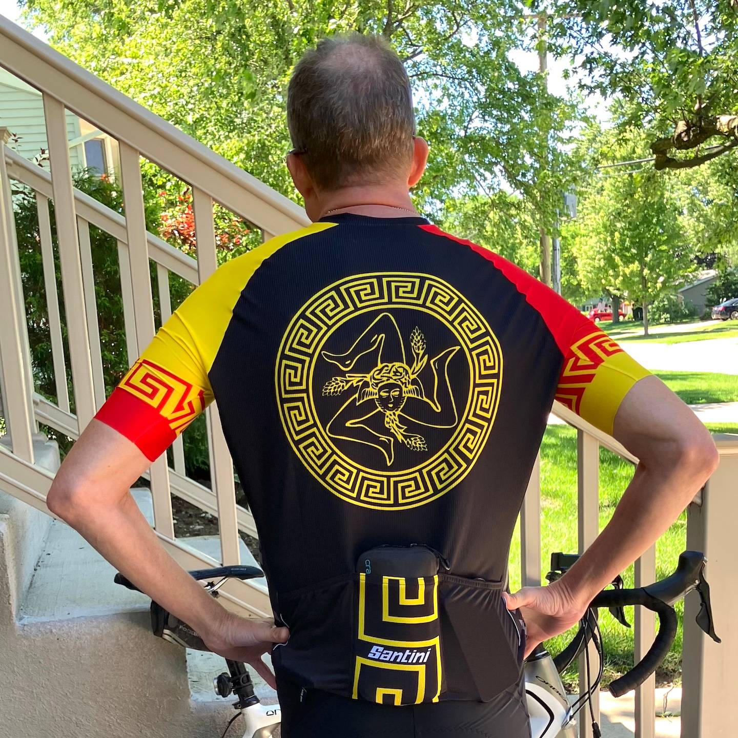 Neil poses in front of the side of front steps in a leafy neighborhood with a red, yellow and white bike jersey showing the circular symbol of Sicily