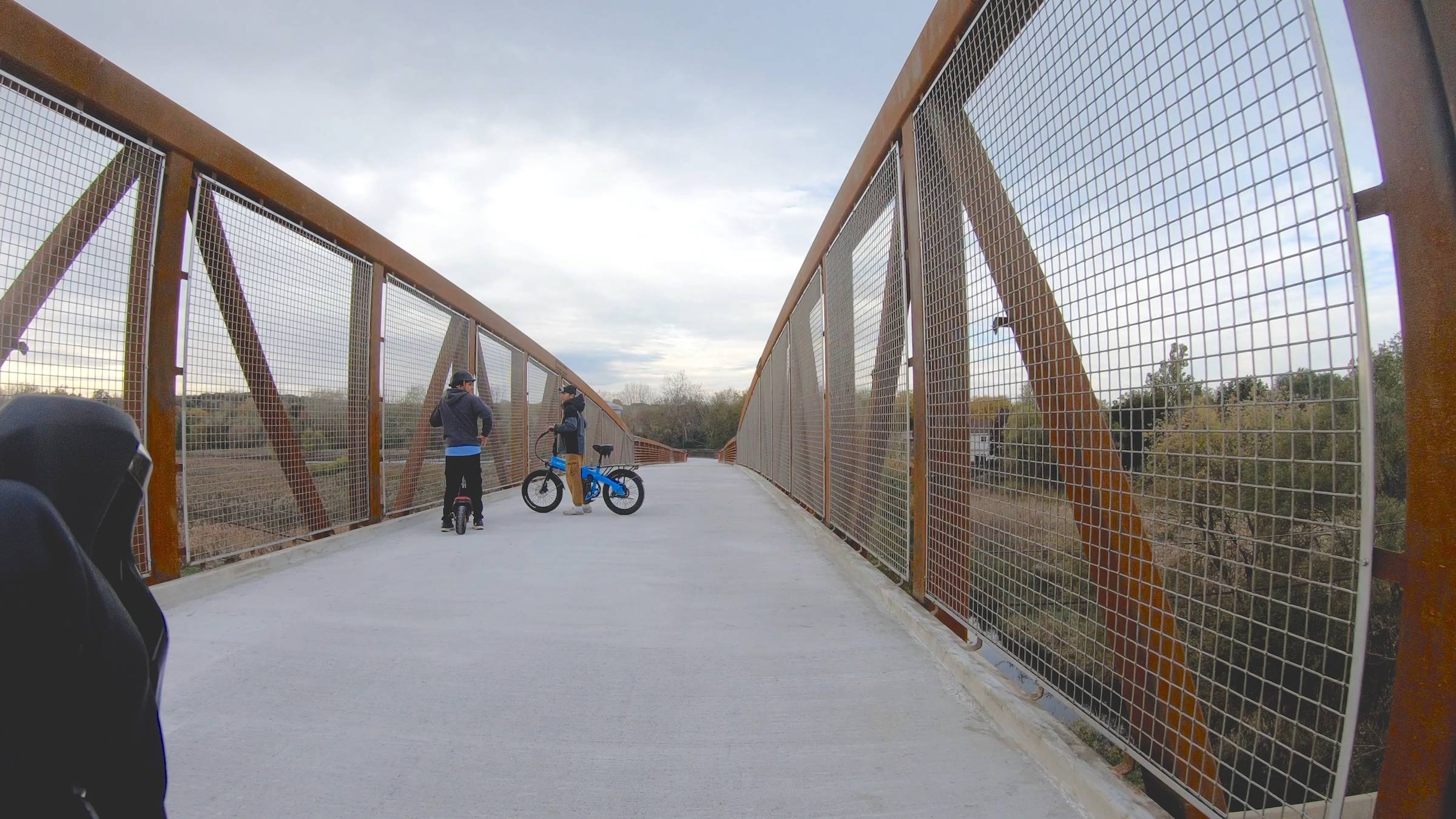 Two boys, one on a scooter and one on an e-bike, stop on a bridge that's near a prairie scene.