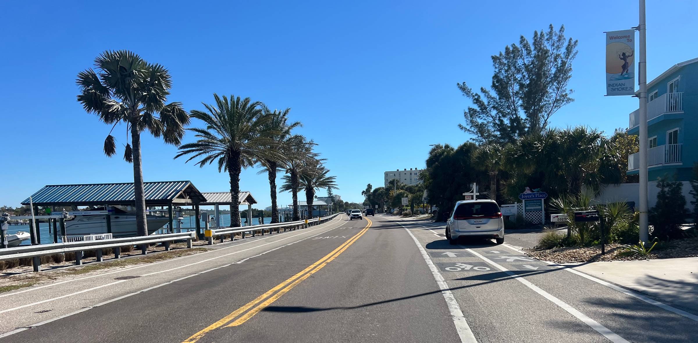 Looking down a two-lane road with bike and pedestrian lanes going by a bay lined with covered boat docks and palm trees.