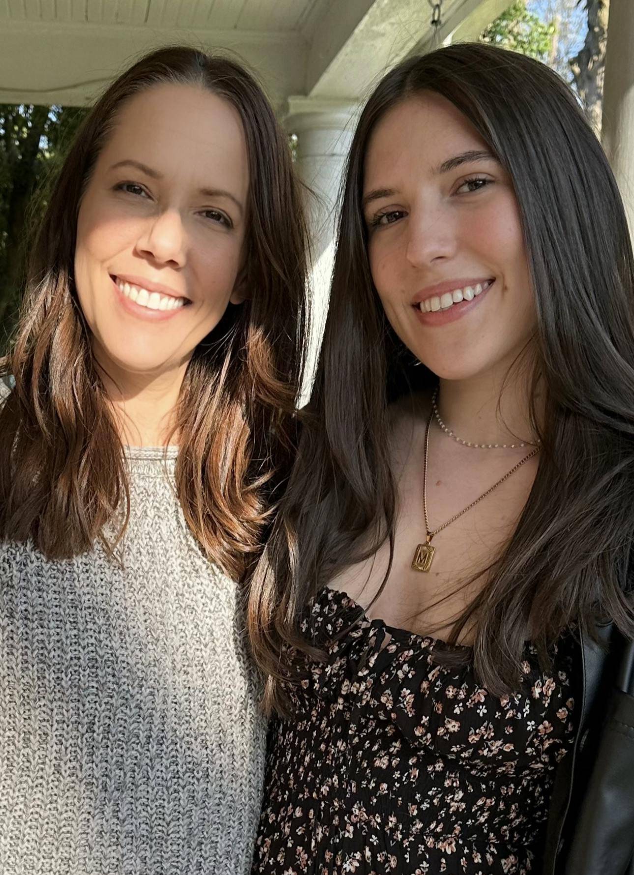 A young woman with long brown hair smiling and standing next to her mother who is also smiling.