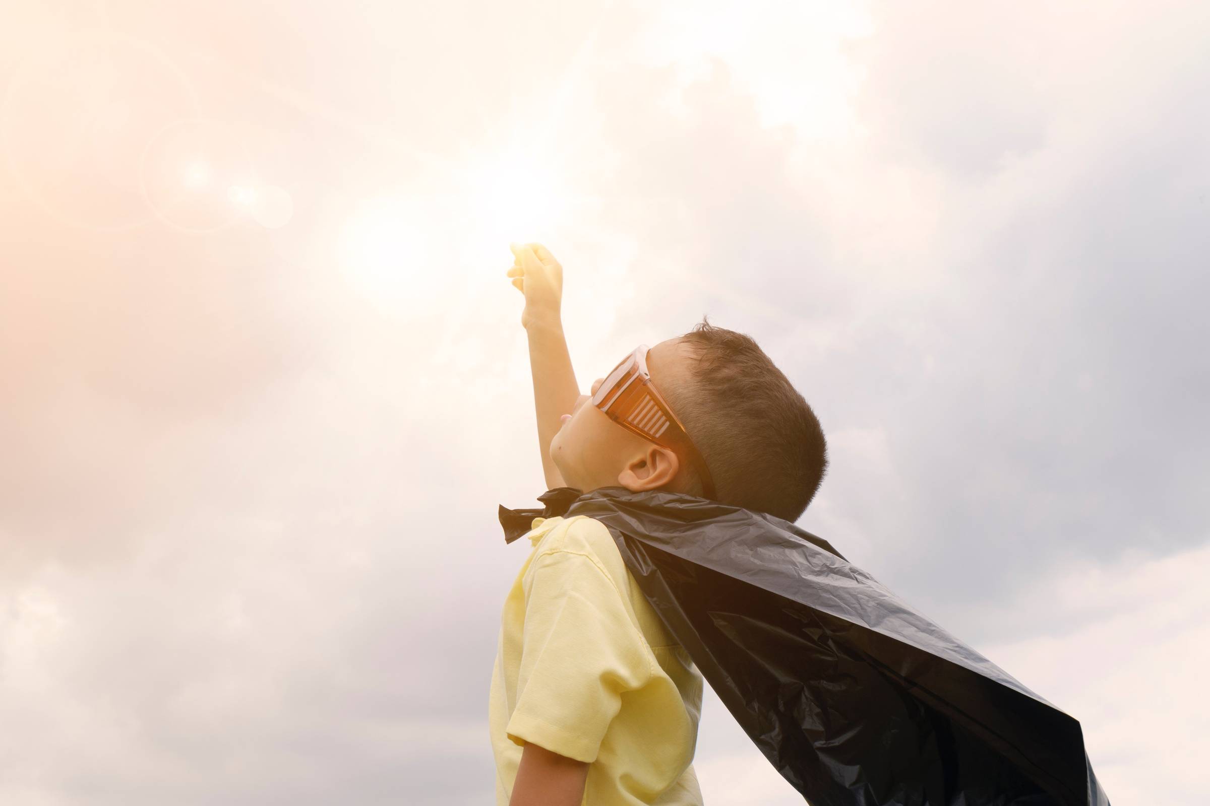 Young boy around age 4-5 dressed up as superhero with a cap and goggles, holding his hand in the air.