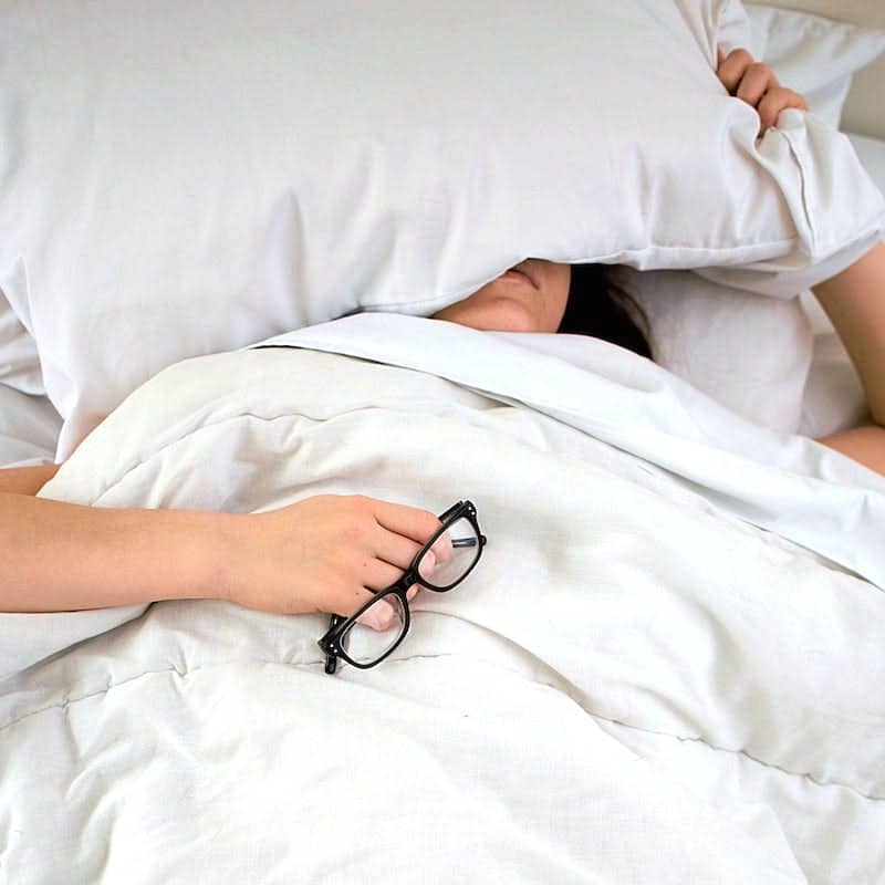 person lying on bed while covering face with pillow and holding eyeglasses