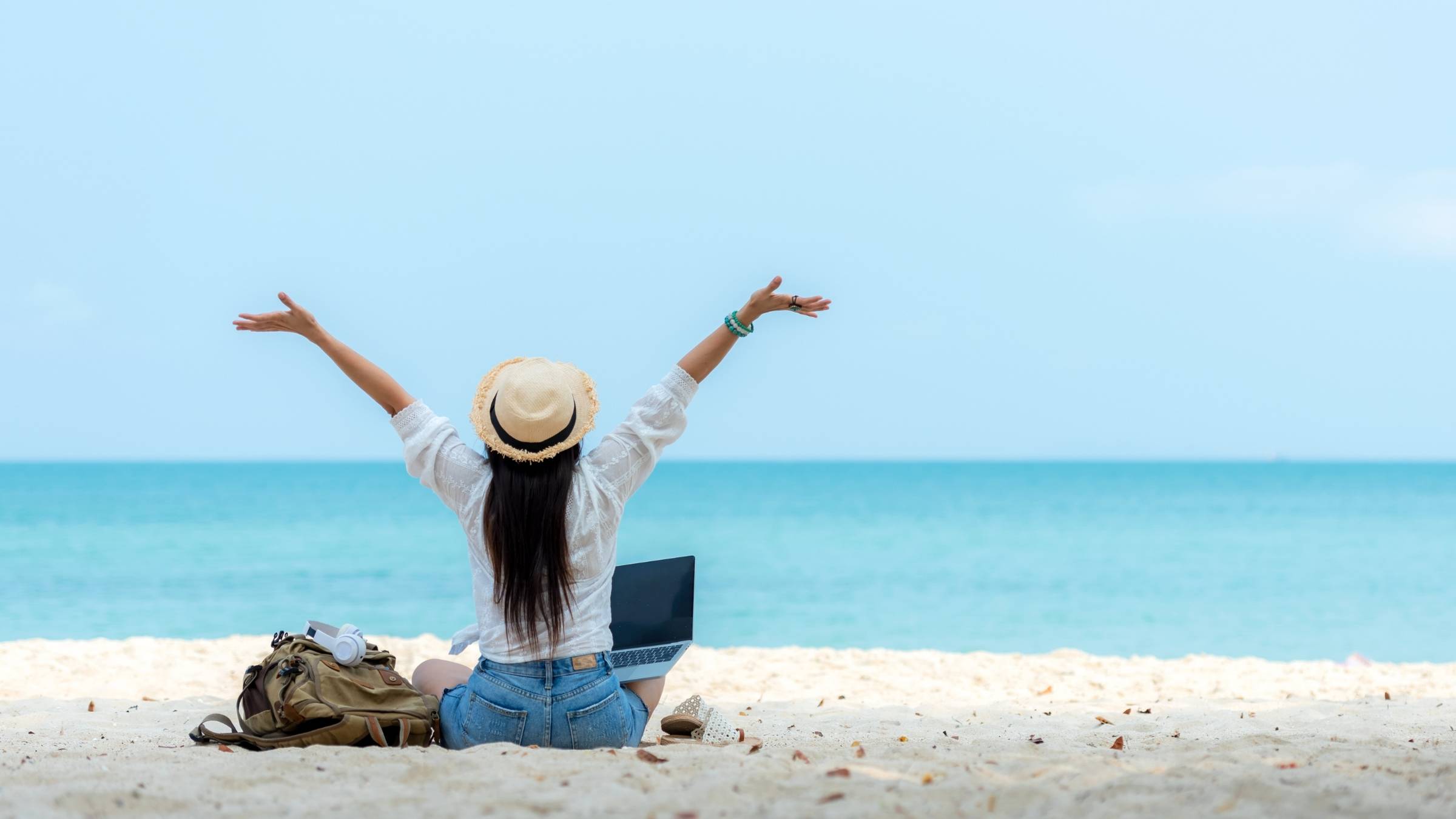Woman on the beach with her arms raised and a laptop beside her