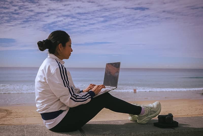 Woman works on laptop by the beach.