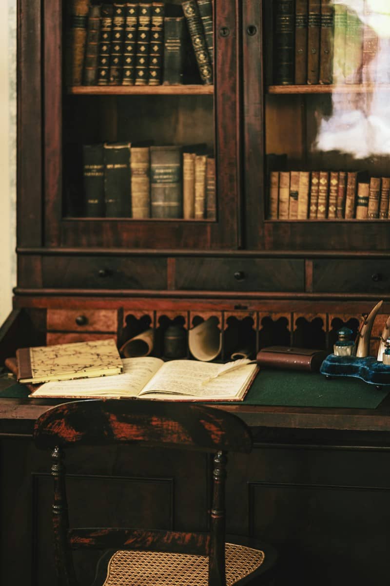 An open book with a quill on a wooden desk before a library cabinet