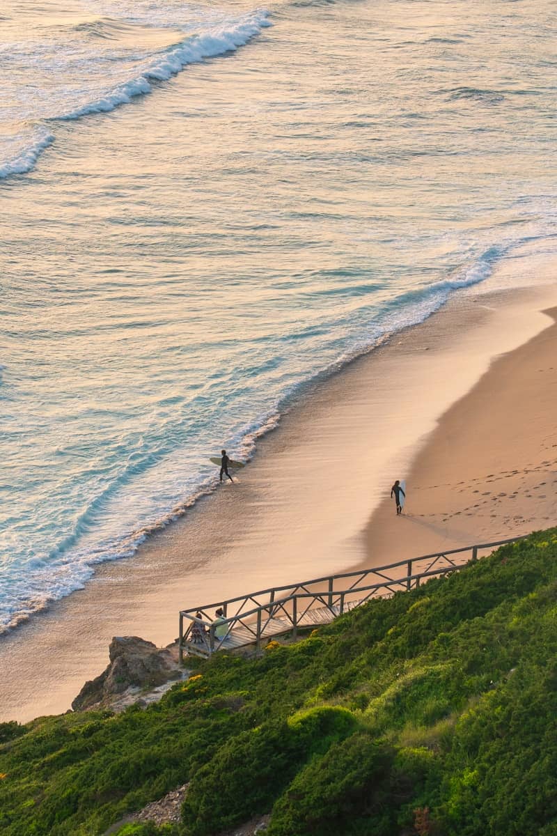 Surfers walk along a peaceful beach at sunset.