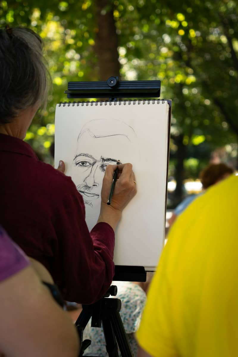 A person in a maroon shirt sketching a portrait on a pad outdoors