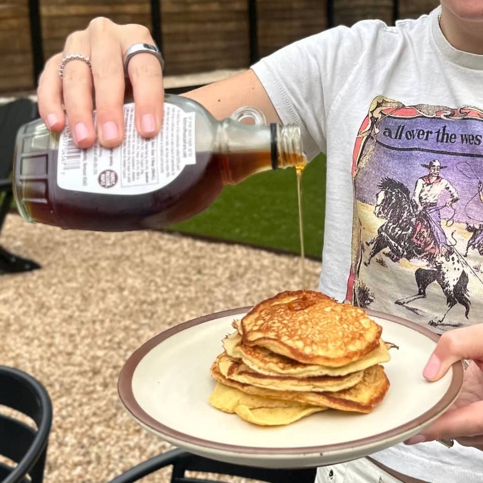 A close-up photo of a person pouring maple syrup from a glass bottle onto a stack of golden-brown pancakes on a beige plate with a brown rim.