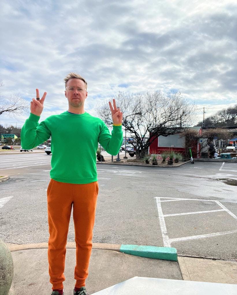 Nick with a green top and orange pants holding peace signs up