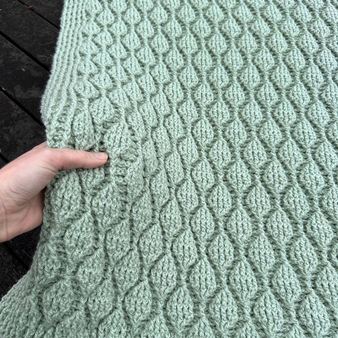A green crochet leaf blanket is laid on a table and a hand is holding it