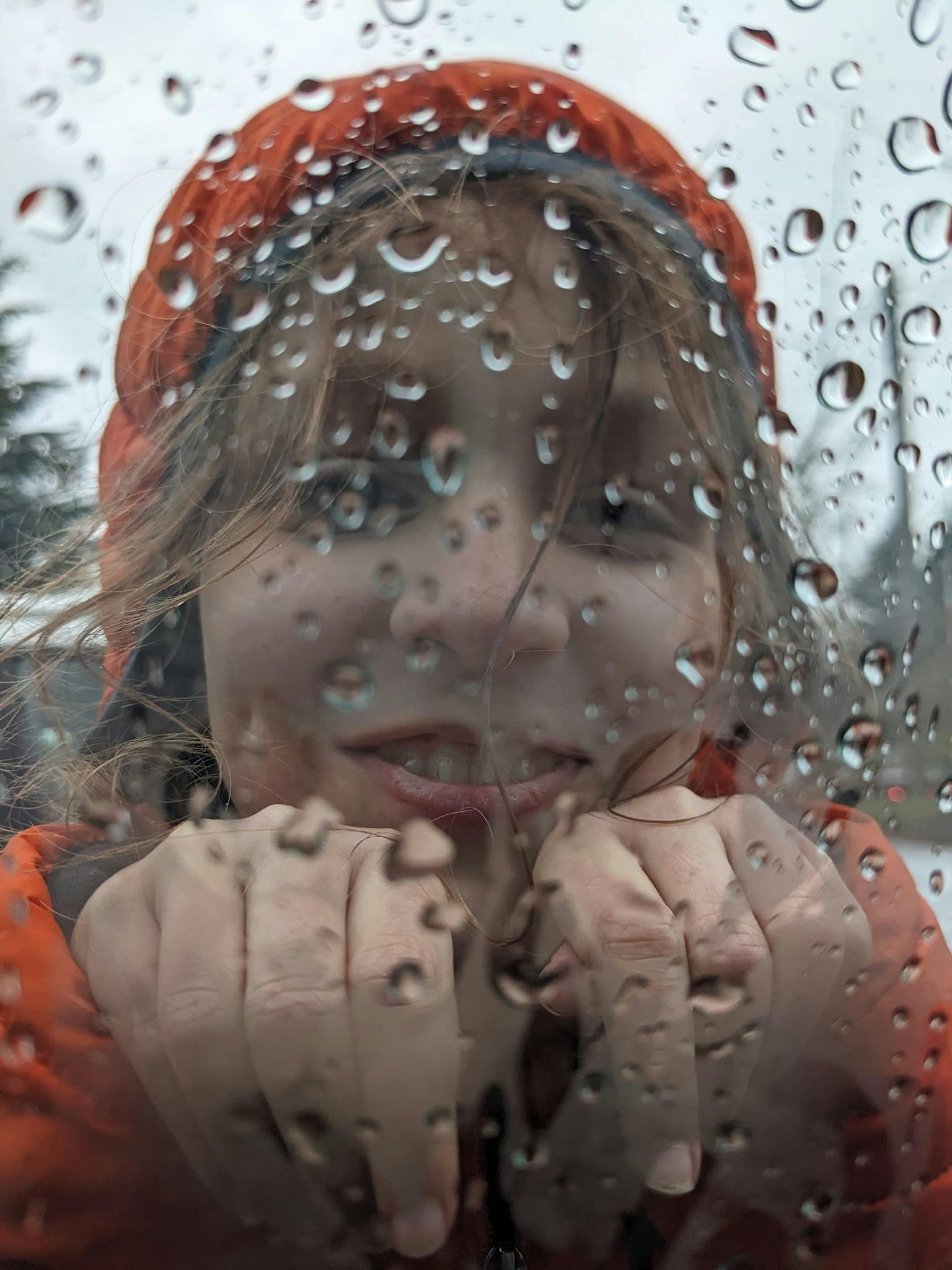 Child looking through a rainy window, wearing an orange coat.