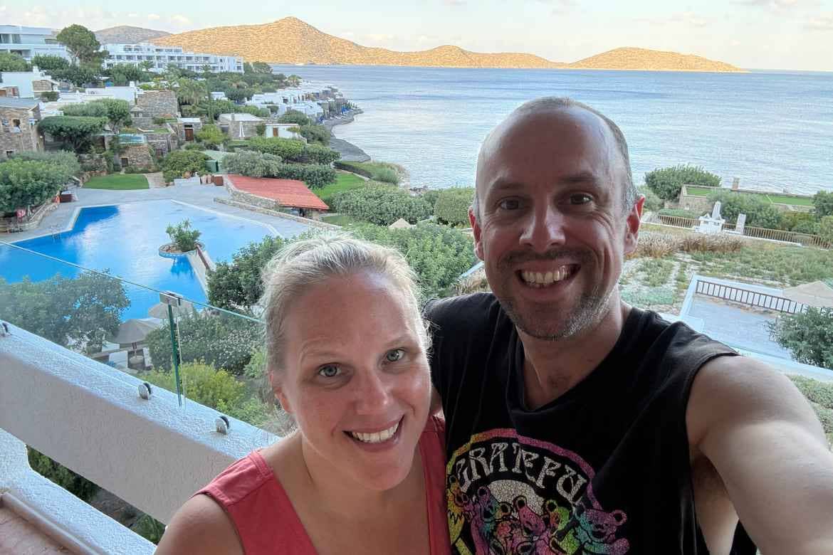 Rachel and Sasha standing on the balcony of their suite at Elounda Mare in Crete with the pool, sea, and mountains in the background