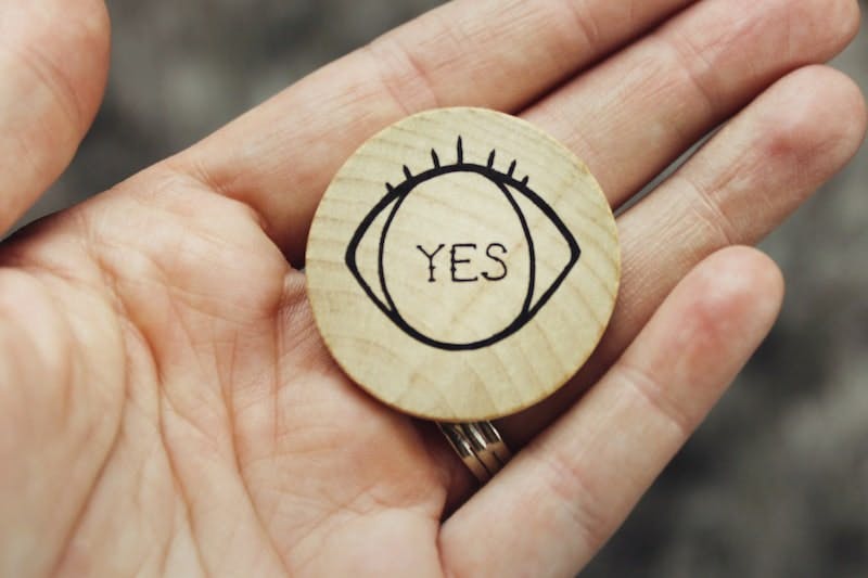 Round wooden coin with eye detail and word, text "yes" printed on it, held in the palm of a woman's hand.
