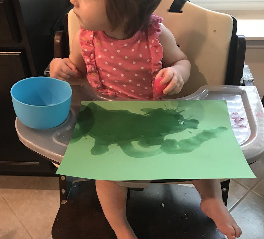 young girl sitting in high chair holding a wet pom pom, blue bowl filled with water and green construction paper that is wet