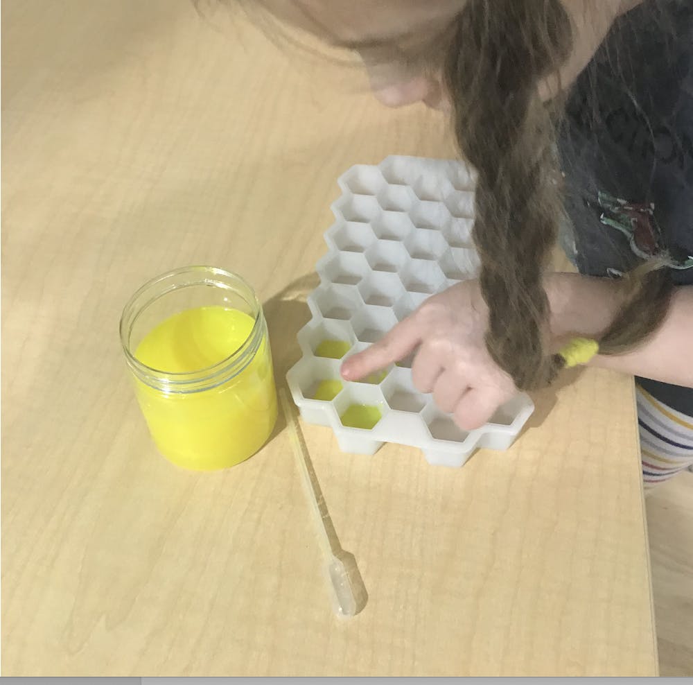young girl pointing at a hexagonal cube tray with a cup of yellow paint and a pipette on a table
