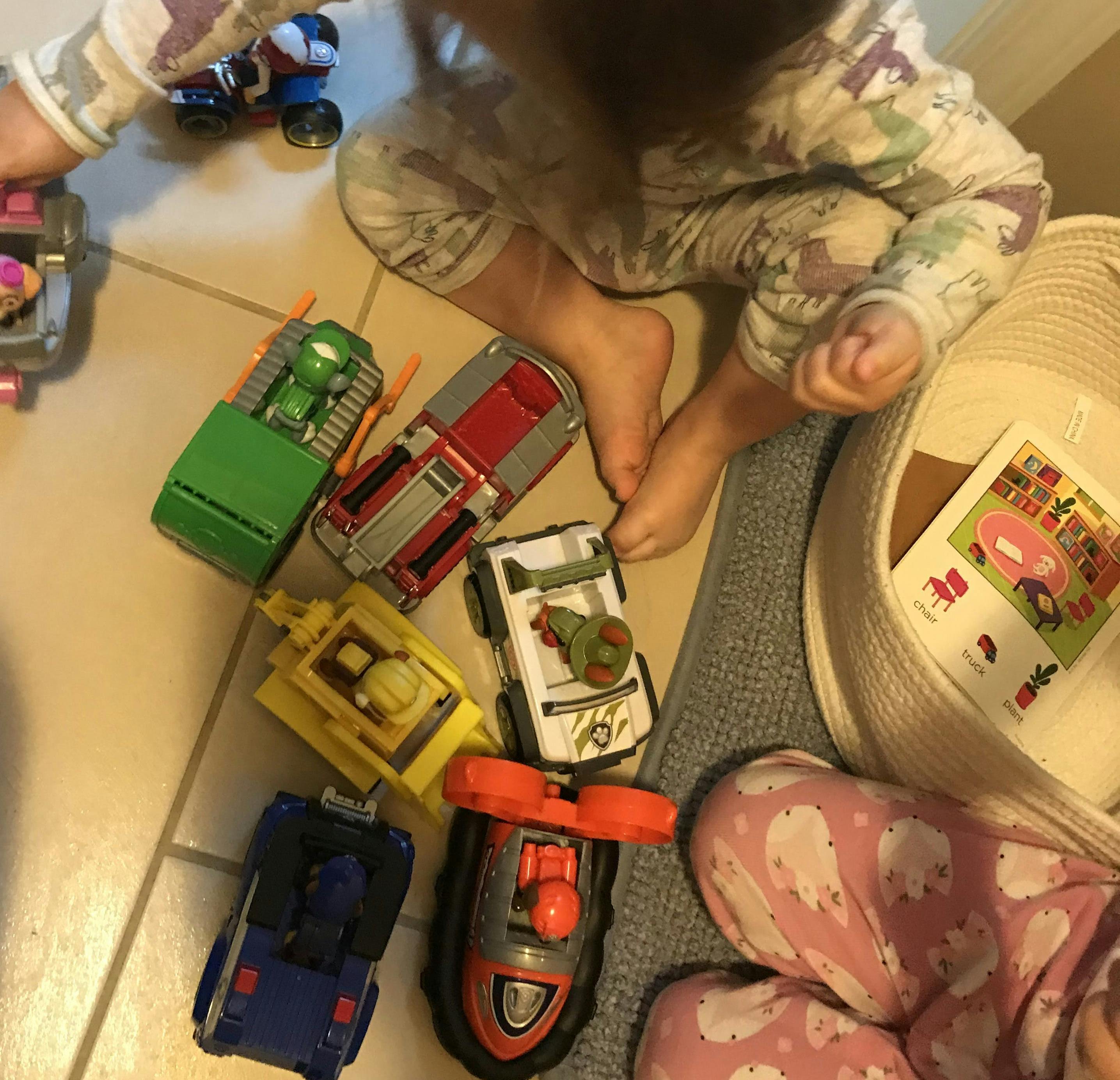 toddler girl playing with Skye from Paw Patrol with all other Paw Patrol toys strewn about, second girl sitting next to a basket with a magazine inside