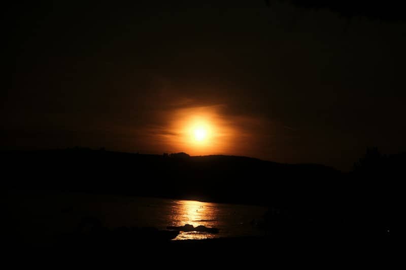 A bright low sun casting a golden reflection on dark water near a hillside