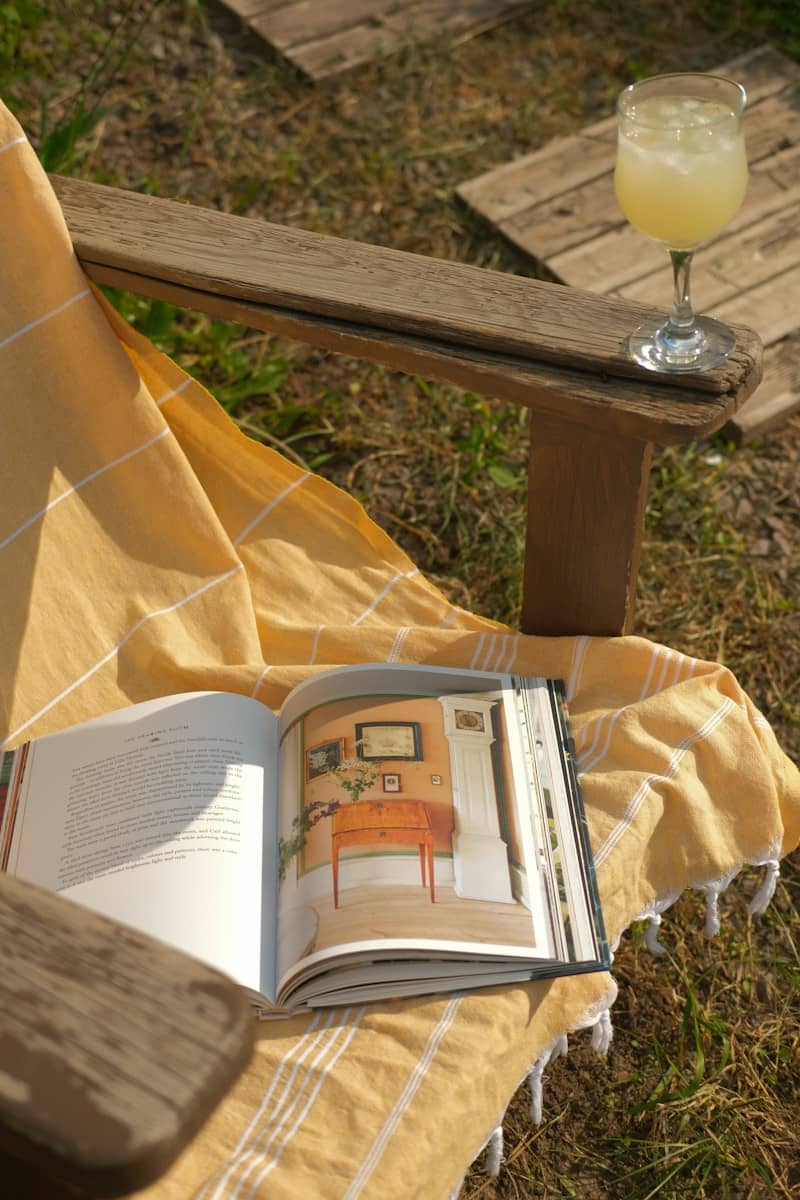 Open book, drink, and yellow blanket on a wooden chair.