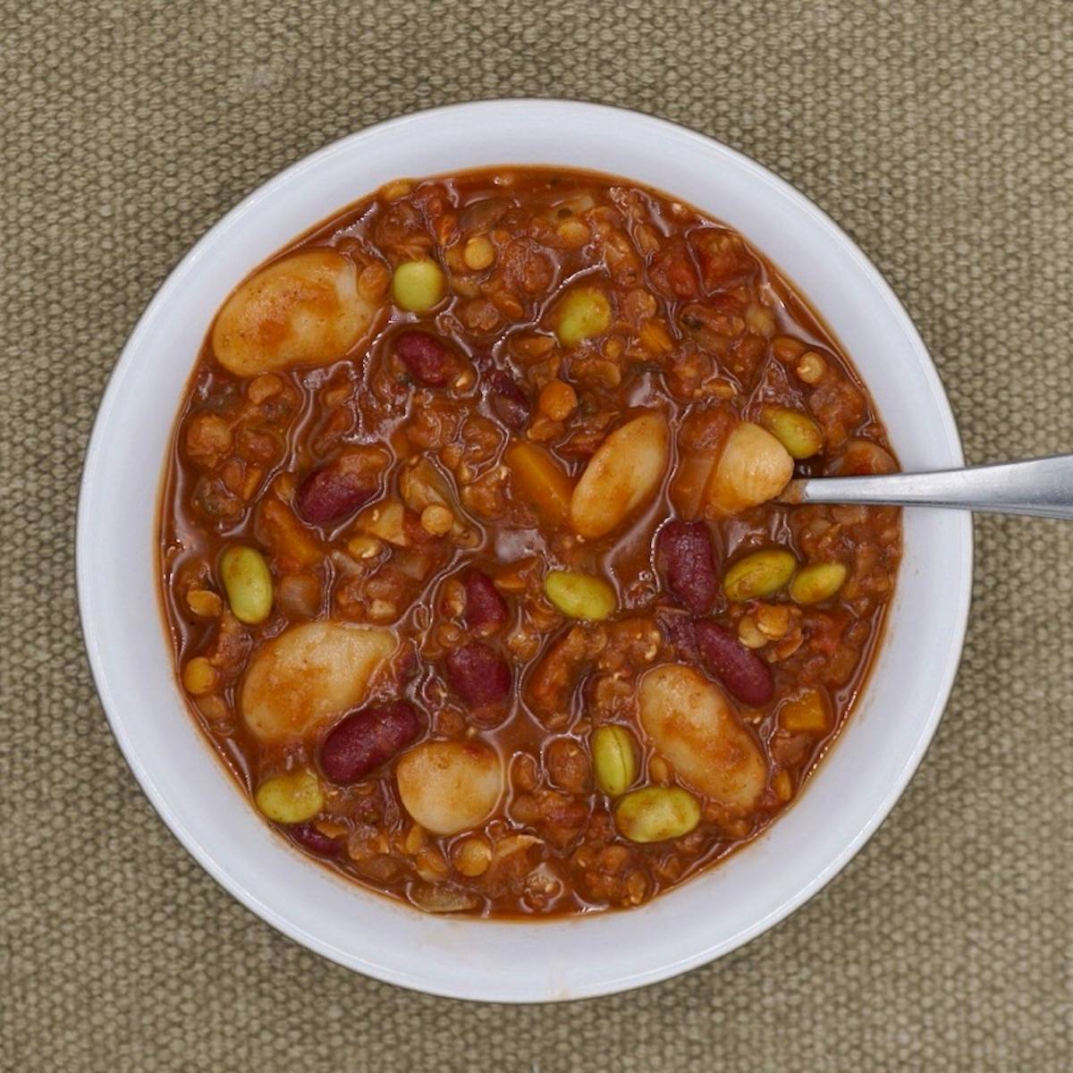 A bowl filled with a hearty bean soup, which includes a variety of beans such as kidney beans and lima beans, in a thick tomato-based broth. A spoon is placed inside the bowl. The bowl is set against a textured, woven fabric background.